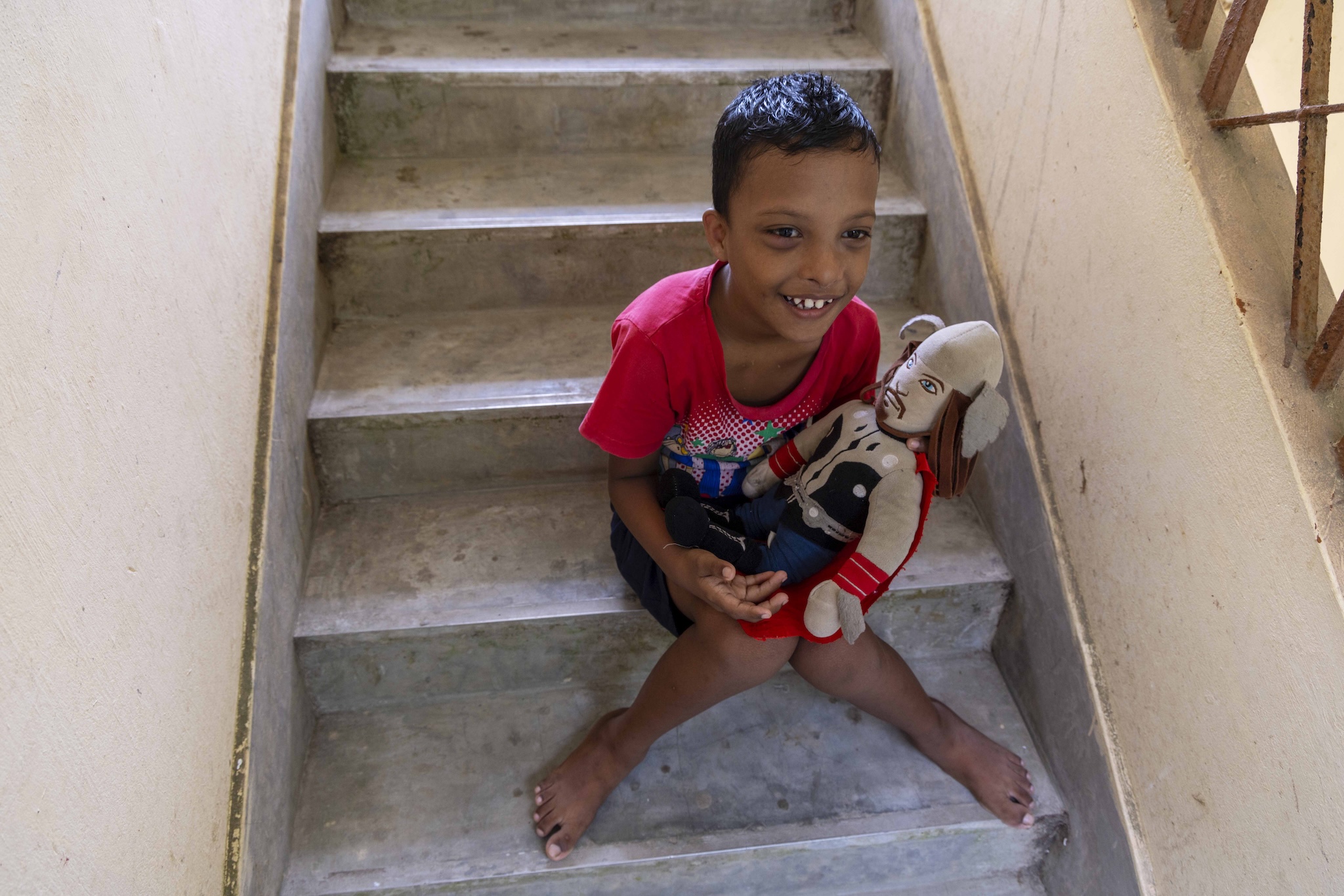 Abhash Minj (10) sits on the grey cement steps of a stairway, smiling as he holds a homemade stuffed cloth doll. He has cropped black hair and wears a red T-shirt and navy blue shorts. His knees touch so that his legs form an inverted V. The doll is the figure of a man made of beige fabric. The features on his face are thin strips of brown cloth that outline eyes, eyebrows, nose, a thin moustache and a horizontal line for a mouth. The pupils of the eyes are made of sky blue cloth. The hair is composed of strips of brown felt material. Wound around the wrists of the stubby arms are three bands of red cloth. There is a large navy blue patch on either side of the chest and a thin vertical navy blue line running down the chest. The stubby legs ‘wear’ steel blue shorts.