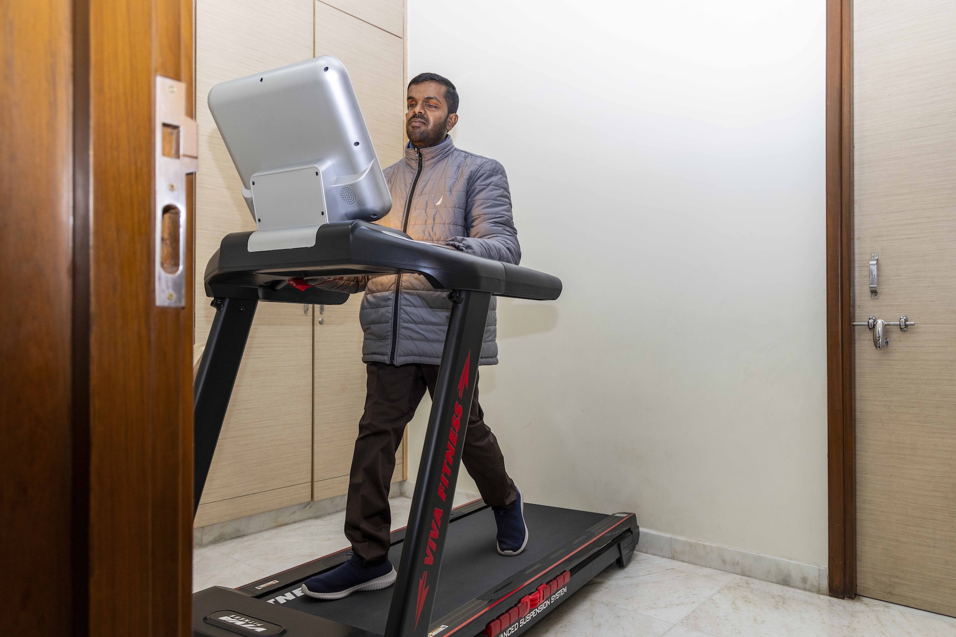 Akshay Surana (34), with eyes closed, is walking on a black treadmill placed in the corner of a room with a white marble floor and off-white walls. He has cropped black hair and closely trimmed moustache and beard. He wears navy blue slip-on shoes, dark brown trousers, and a light grey puffer jacket. Both his hands are on the horizontal bar of the treadmill.