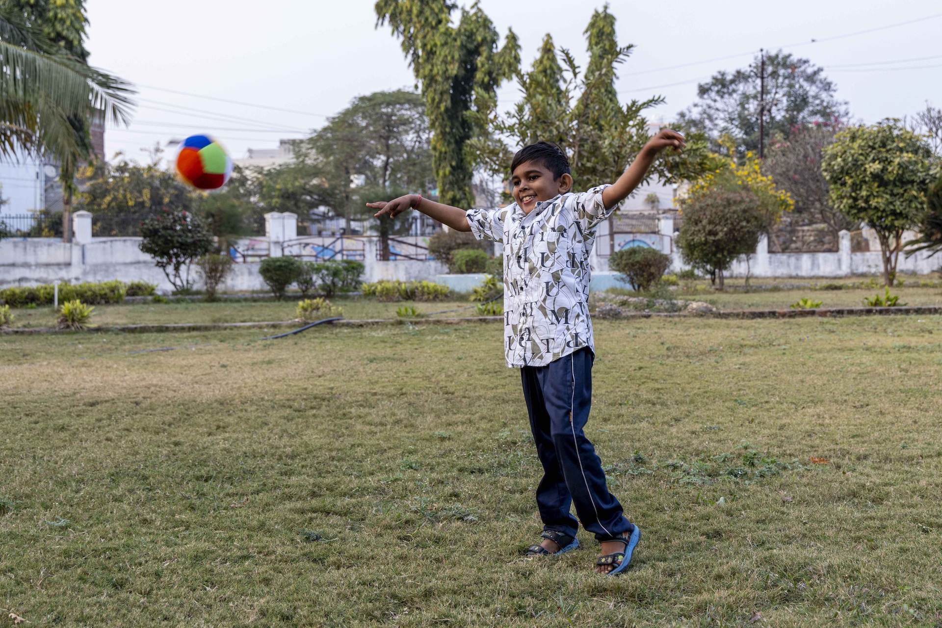 Aman Kumar Nitinbhai (9) stands outdoors on an expanse of stubbly green grass. He has short, straight, black hair combed towards the forehead. He wears navy blue track pants, turquoise blue sandals with ash-grey straps, and a white half-sleeved shirt printed with large, black, random capital letters of the English alphabet and irregular-shaped, pastel-brown patches. He smiles as he raises both hands so they look like wings. His shoulders are shrugged and his right heel is raised while he stands on the toes of his left foot. It appears as if he has just thrown a ball because a soft ball made of plush is suspended mid-air in front of him. The ball has sections in a hexagonal honeycomb pattern, and the side facing the camera are coloured red, sky blue, and sapphire blue. In the far distance there is a whitewashed compound wall and green trees and shrubs.