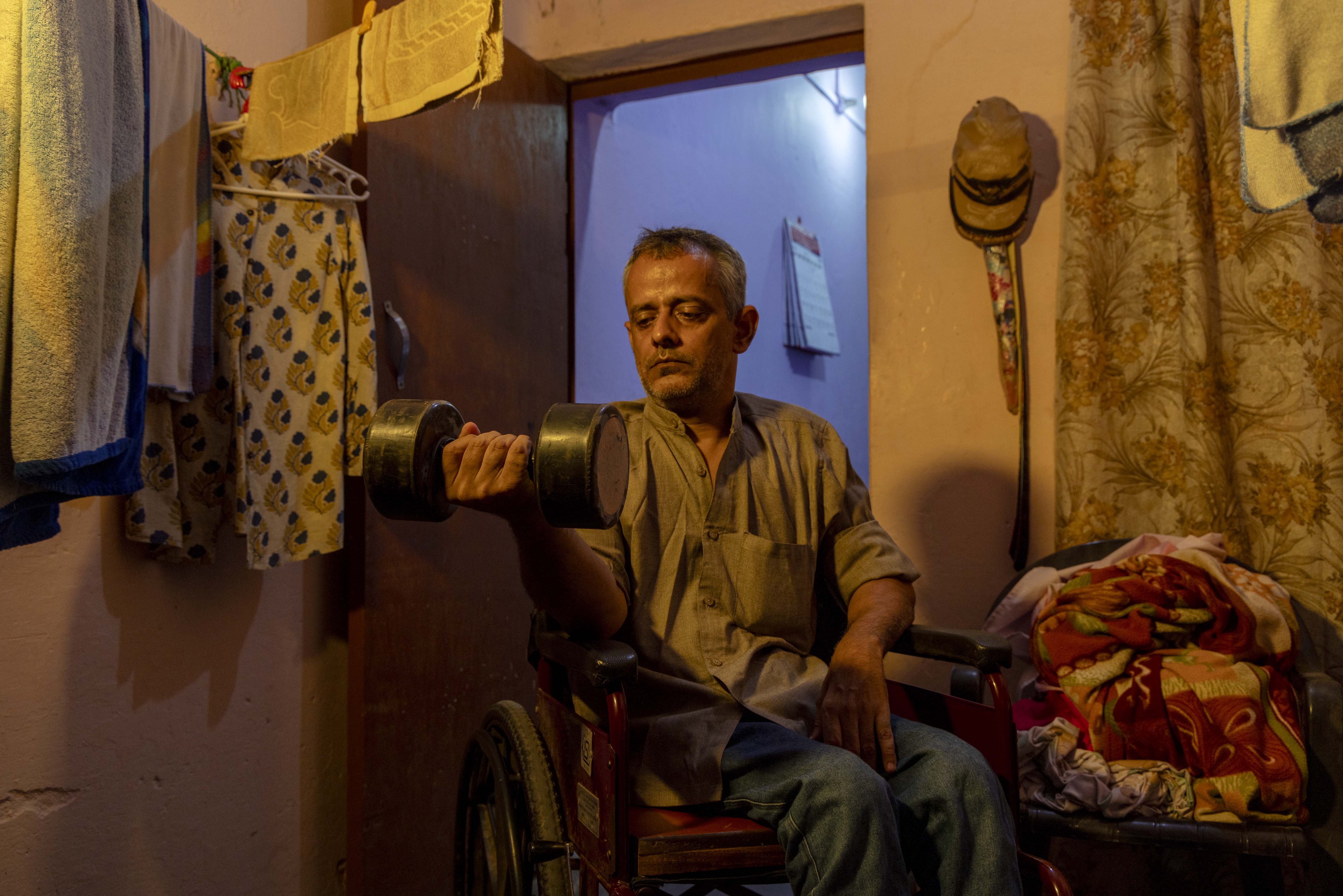 Amit Bhatla (50) sits in a brown wheelchair in a room with soft yellow lighting. He wears a snuff-brown half-sleeved shirt with a Chinese collar and light blue jeans. He has a stubbly crop of salt-and-pepper hair and an equally stubbly beard and moustache. He is lifting a black dumbbell with his right hand while his left hand lies on the left armrest. The room’s walls are salmon-pink. The dark brown door behind him is ajar, revealing the light blue wall of the adjacent room.