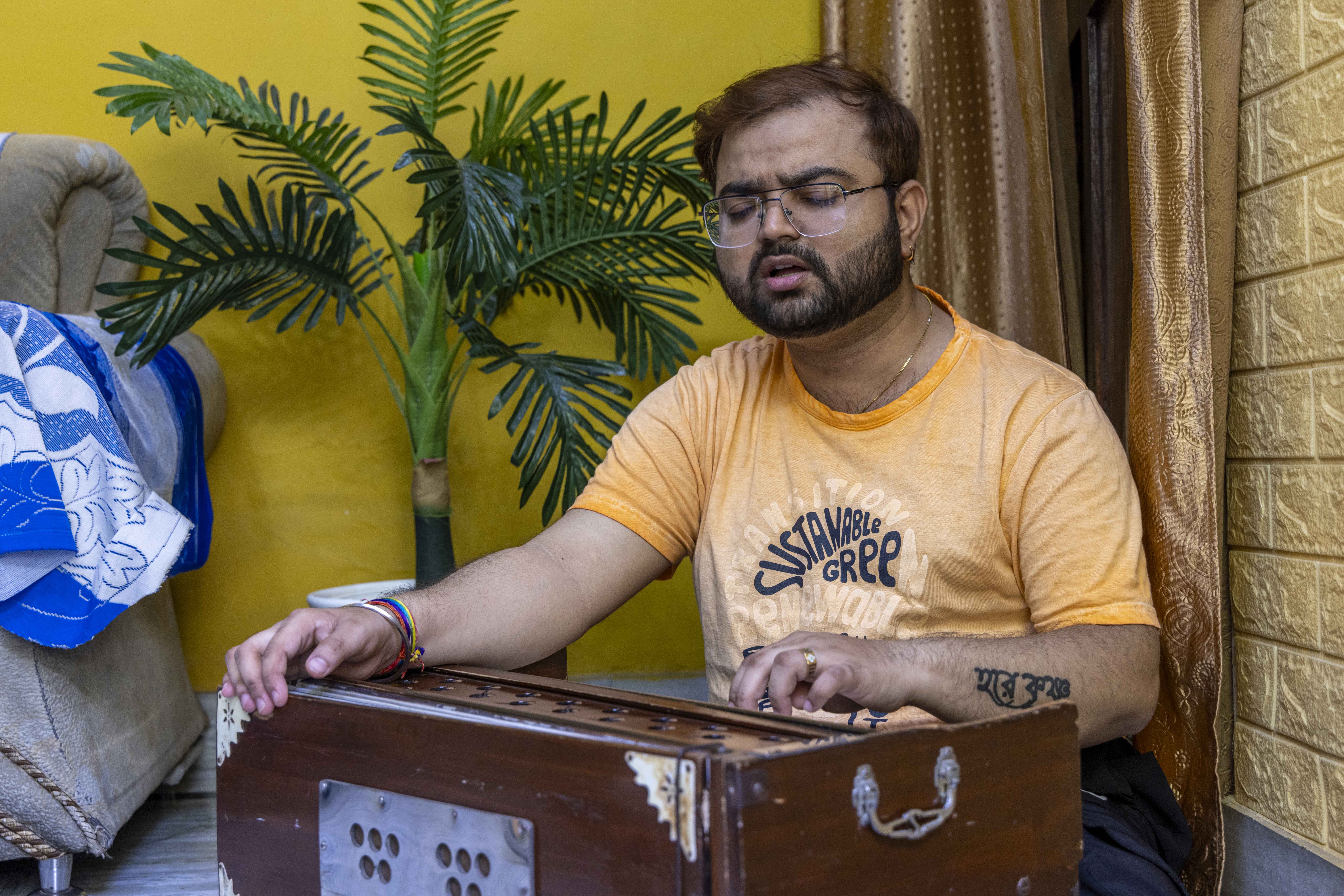 Amit Kumar (34) sits in a corner of a room on the floor behind a chocolate brown harmonium. He wears rimless glasses and his short, light brown hair has a side parting. His black moustache curves down to meet his thin black beard that reaches his ears. He wears a pale orange T-shirt with black lettering on the chest, and black trousers. His mouth is slightly open and his right hand is on the bellows while his left hand plays the keys. The wall behind him is mustard-yellow and there is a short green ornamental palm tree in front of it.