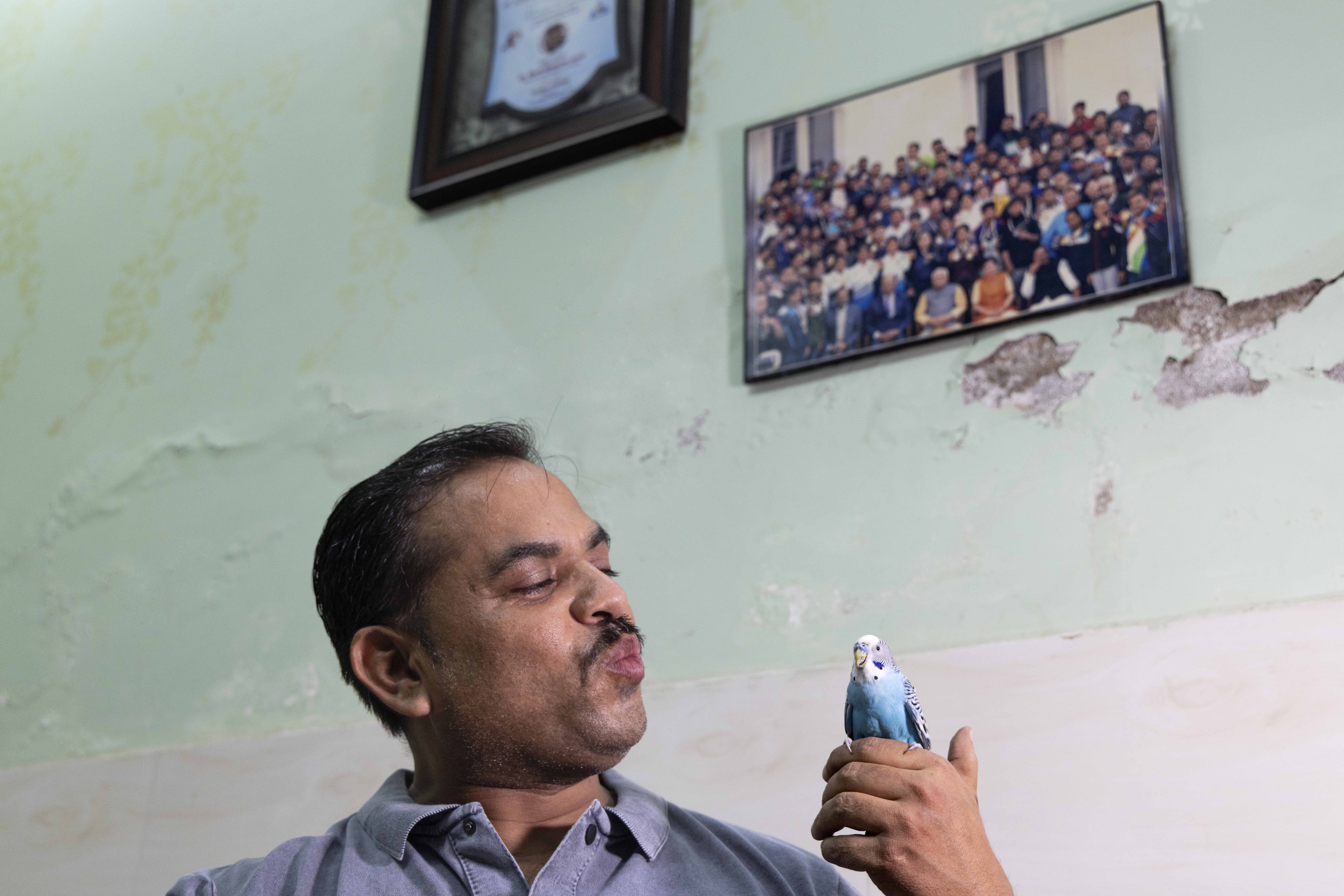 Close-up of Amresh Kumar Singh (44) looking at a lovebird he holds up in his left hand. He has a thin moustache and cropped hair and wears a light blue shirt. The bird is sky-blue with a white head. Amresh has puckered his lips as if to kiss the bird. The wall behind them is a faded pista green and there is a framed group photo hanging on it. The photo shows blurry faces of 30 or 40 people seated in rows.