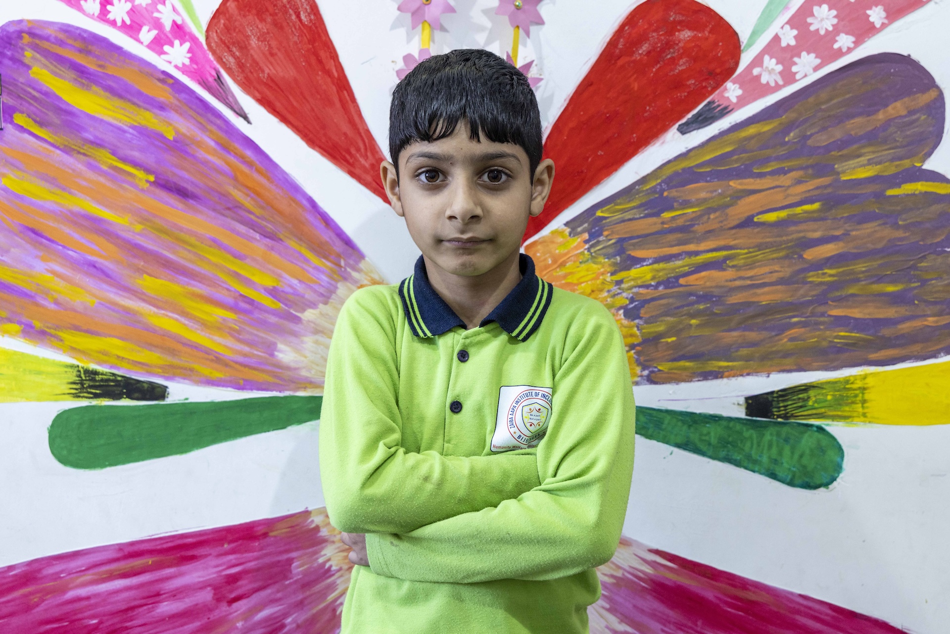 Mid shot of Arhan Khursheed (8) standing with folded arms. He wears a lime-green, full-sleeved knitted shirt with a black collar. His cropped, straight, black hair is combed forward to his forehead. The wall behind him is covered with huge club-shaped murals coloured pink, green, red, yellow, and purple. The club shapes fan out as if to form a giant flower.
