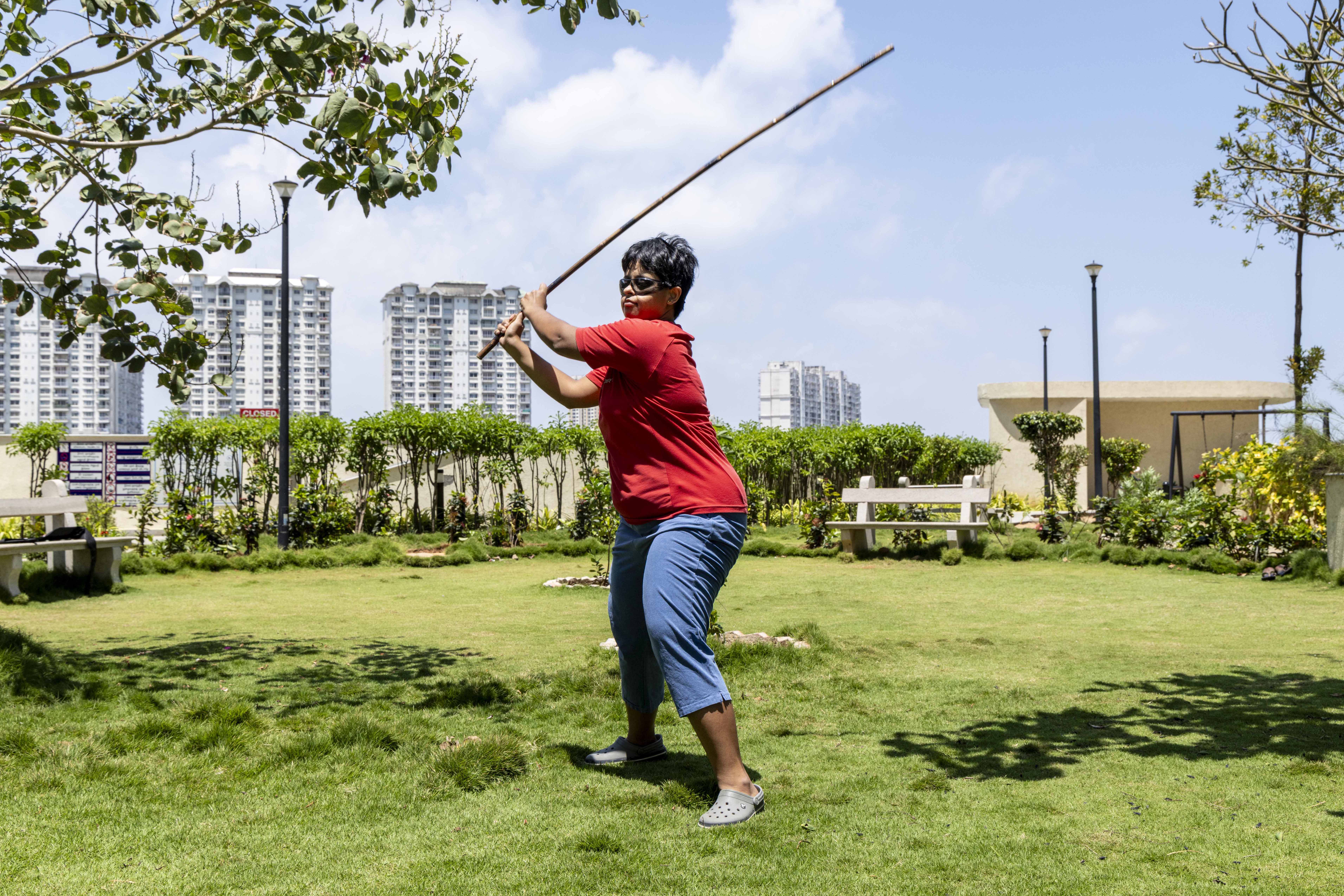 Arti Krishnamoorthy (36) stands on a green lawn fringed by green bushes at the perimeter. She has a cropped straight black hair with a side parting and wears dark glasses. She is dressed in a bright red T-shirt and knee-length light blue jeans and wears grey Crocs on her feet. She holds aloft a long, thin wooden rod with both hands while bending her left knee slightly and supporting her weight on the right foot. In the distance there are several multi-storeyed apartment blocks painted white, and a pale blue sky with puffy white clouds.