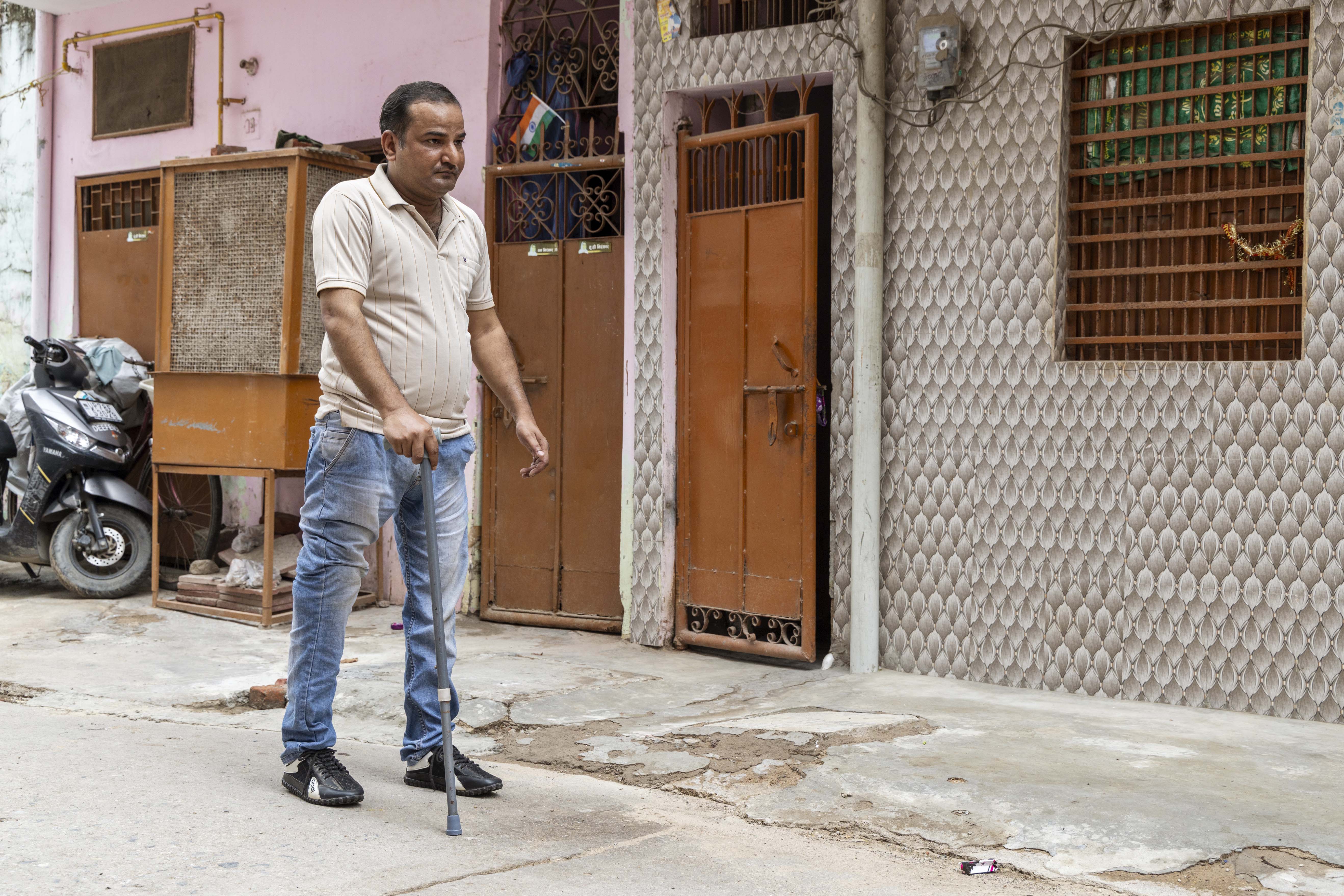 Arvind Kumar (41) stands outside on a roughly paved grey road. He is clean-shaven and has a close crop of black hair. He wears a cream, half-sleeved, knitted shirt, faded blue jeans, and black sports shoes. He holds a grey walking stick in his right hand. The outer wall of the house behind him is covered with tiles in a light brown oval pattern. The wall has a window with iron grills and a metal door, both painted tan brown. The wall is joined to the light pink wall of another house with a similar brown metal door.