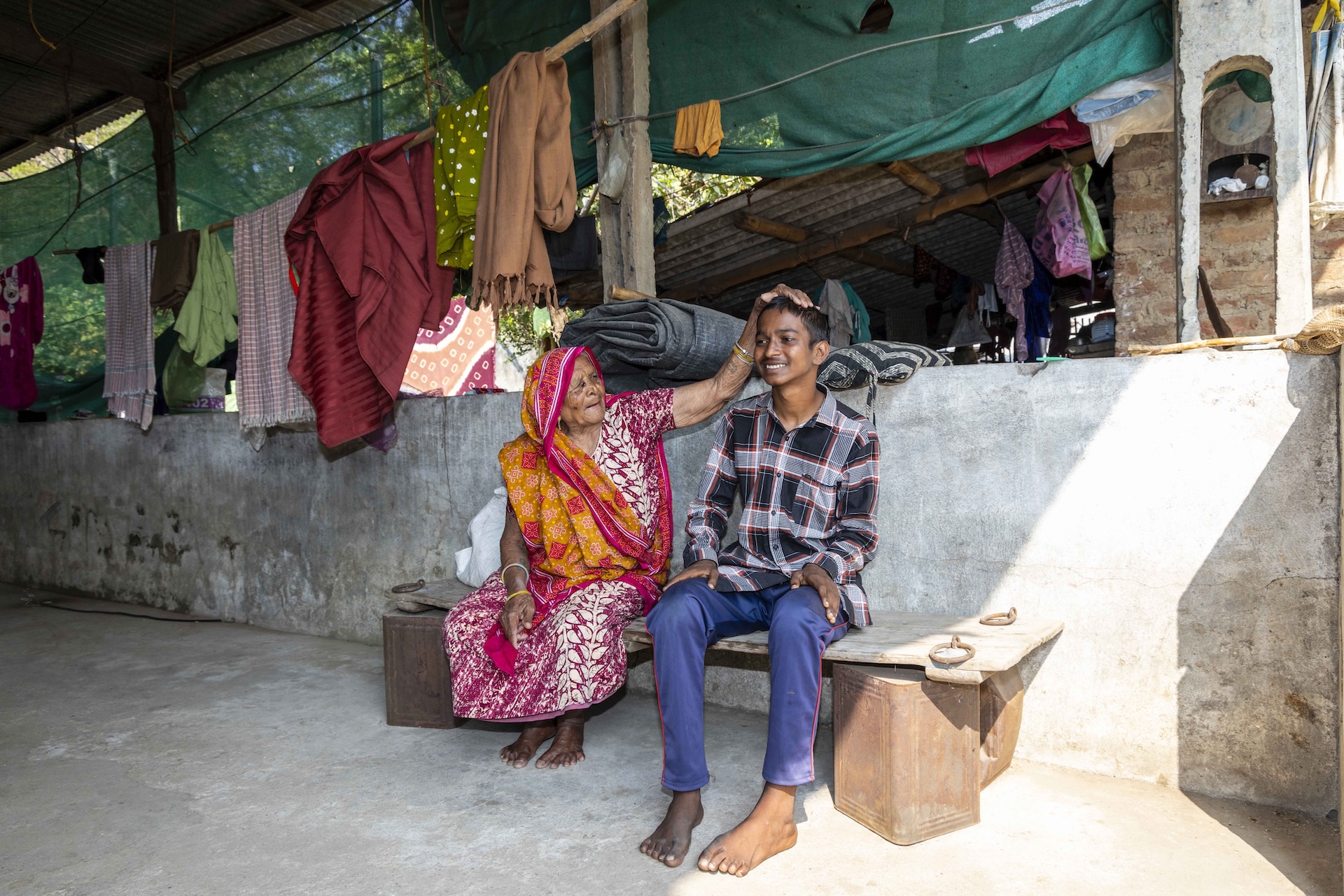 Axay Kumar (16) sits next to his grandmother on a bench made out of a wooden slab resting on two large wooden blocks. He has closely cropped hair combed forward towards his forehead. He wears navy blue track pants paired with a full-sleeved shirt in a chequered pattern of rust-red, white and black. He is smiling as his grandmother Keshaliben (71) turns to place her left hand on top of his head. Keshaliben wears a half-sleeved, round-necked cotton nighty in a red and white design. Draped over her head and shoulders is a dupatta in a tie-and-dye design of red, white and orangish-brown. The floor is plain grey cement and a low cement wall runs the length of the space. The bench touches the wall.