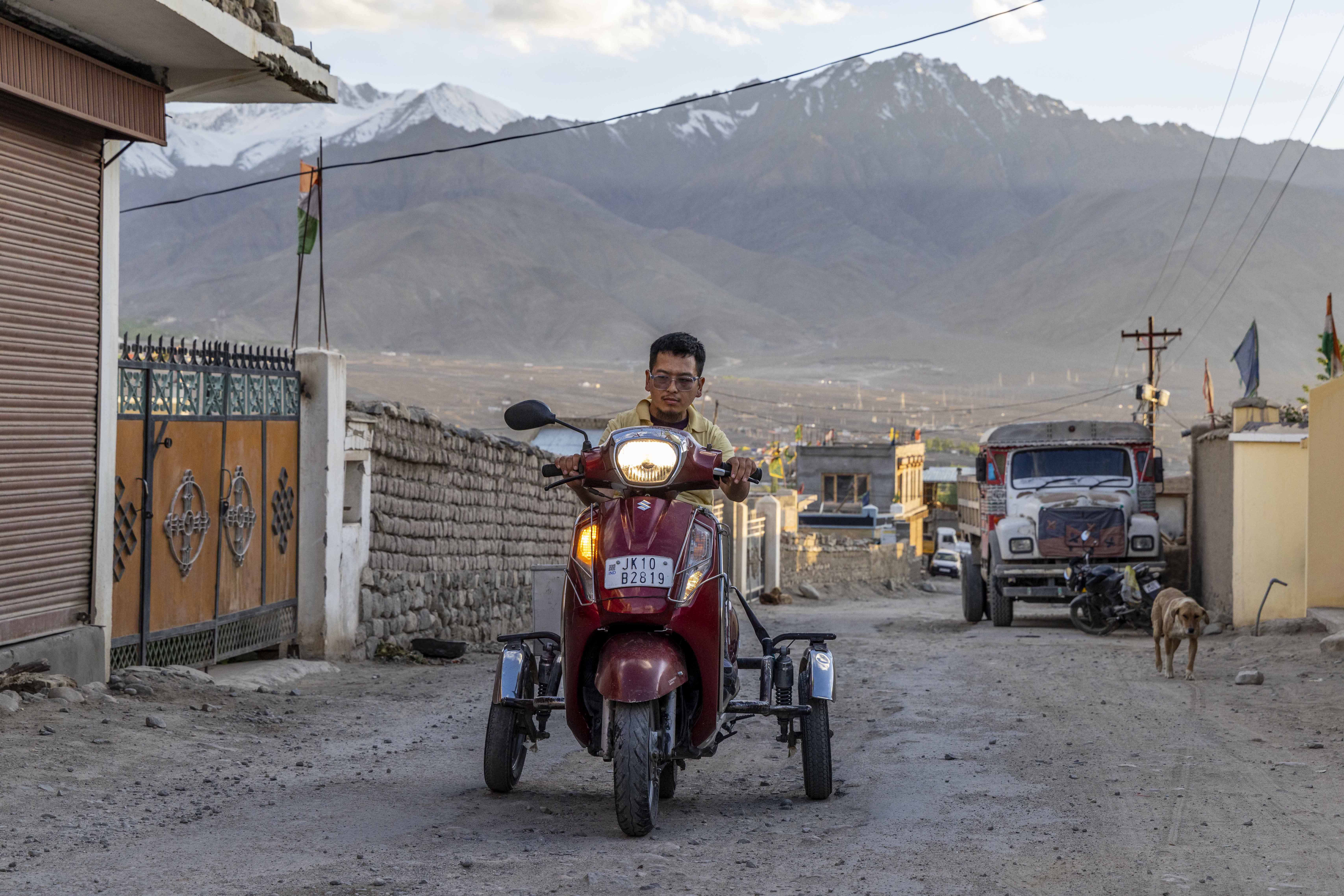 Chemet Rafail (24) sits on a maroon disability scooter facing the camera. The scooter’s headlamp is burning and it has a single black rear view mirror. Rafail has cropped black hair and wears specs and a cream half-sleeved knitted shirt with collar. Behind him a range of mountains looms, with white snow capping the tops of a few ridges. A broad, uneven, unpaved, grey road slopes downward behind him. To the left of the scooter there is a tan-coloured metal gate topped with black iron spikes. A wall next to the gate is made of brown and grey stones and it stretches downward along the road. On the road, some distance behind the scooter, a white lorry is parked to the right, with a motorbike parked just in front of its bumper.