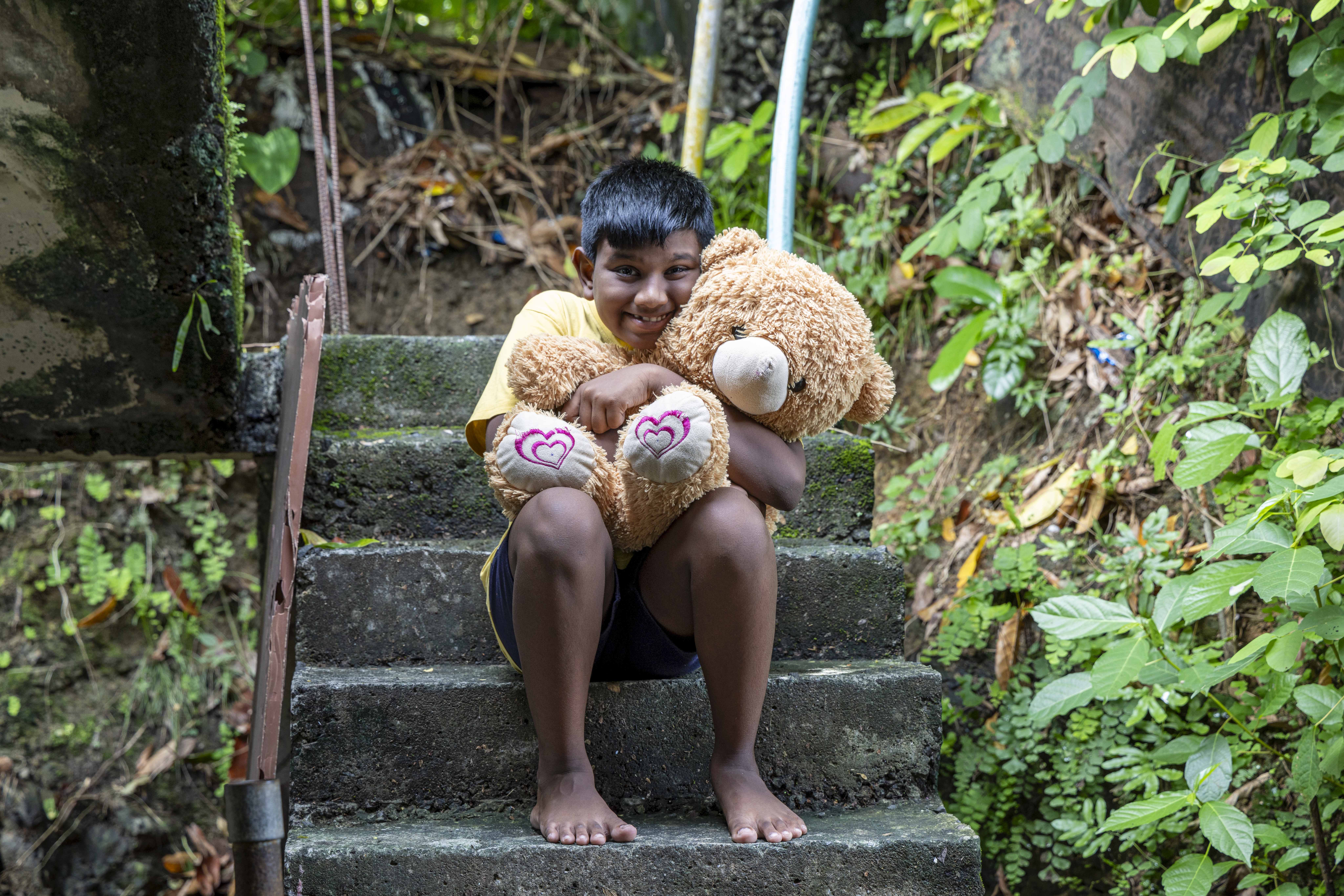 D. Ronak (11) sits sitting outdoors on a flight of plain, dark grey, concrete steps, smiling as he hugs a large sandalwood-coloured teddy bear close to his chest. He has short, straight, black hair combed forward to reach his forehead. He wears a pale yellow T-shirt and black shorts. There is green foliage on either side of the steps.
