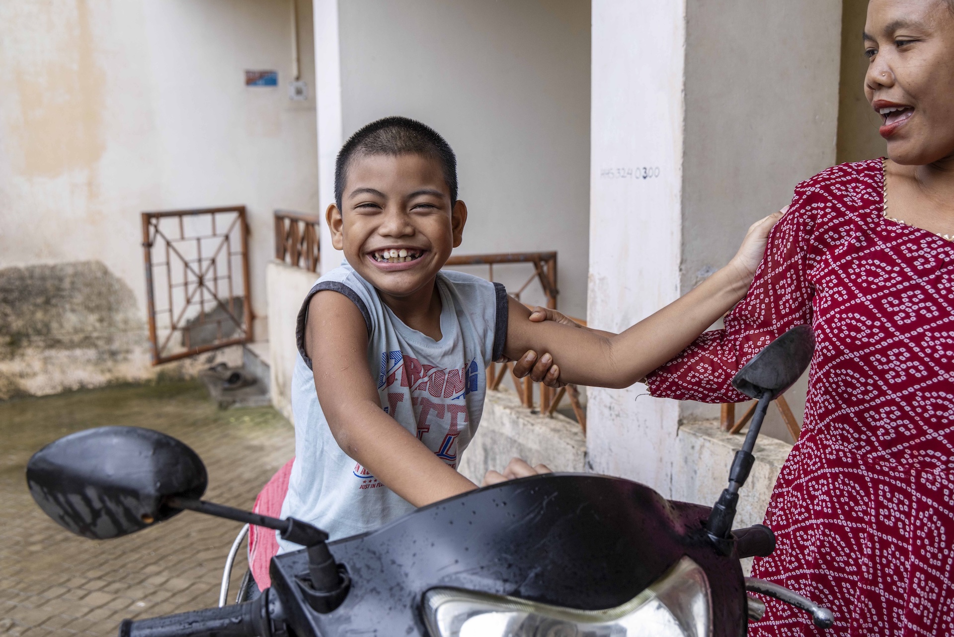 Elation Sultan (9) grins broadly as he sits on a black scooter facing the camera. He has black, closely cropped, stubbly hair. The bottom half of his body is hidden by the scooter’s handlebar. He wears a pale blue sleeveless cotton banian. His right hand presses against the rear of the handlebar and his left hand holds his mother’s shoulder. His mother Eleaner (36) grips his left upper arm with her right hand as she turns sideways to look at him. She wears a three-fourth-sleeved, square-necked, scarlet maxi dress with a white bandhani (tie-and-dye) pattern.