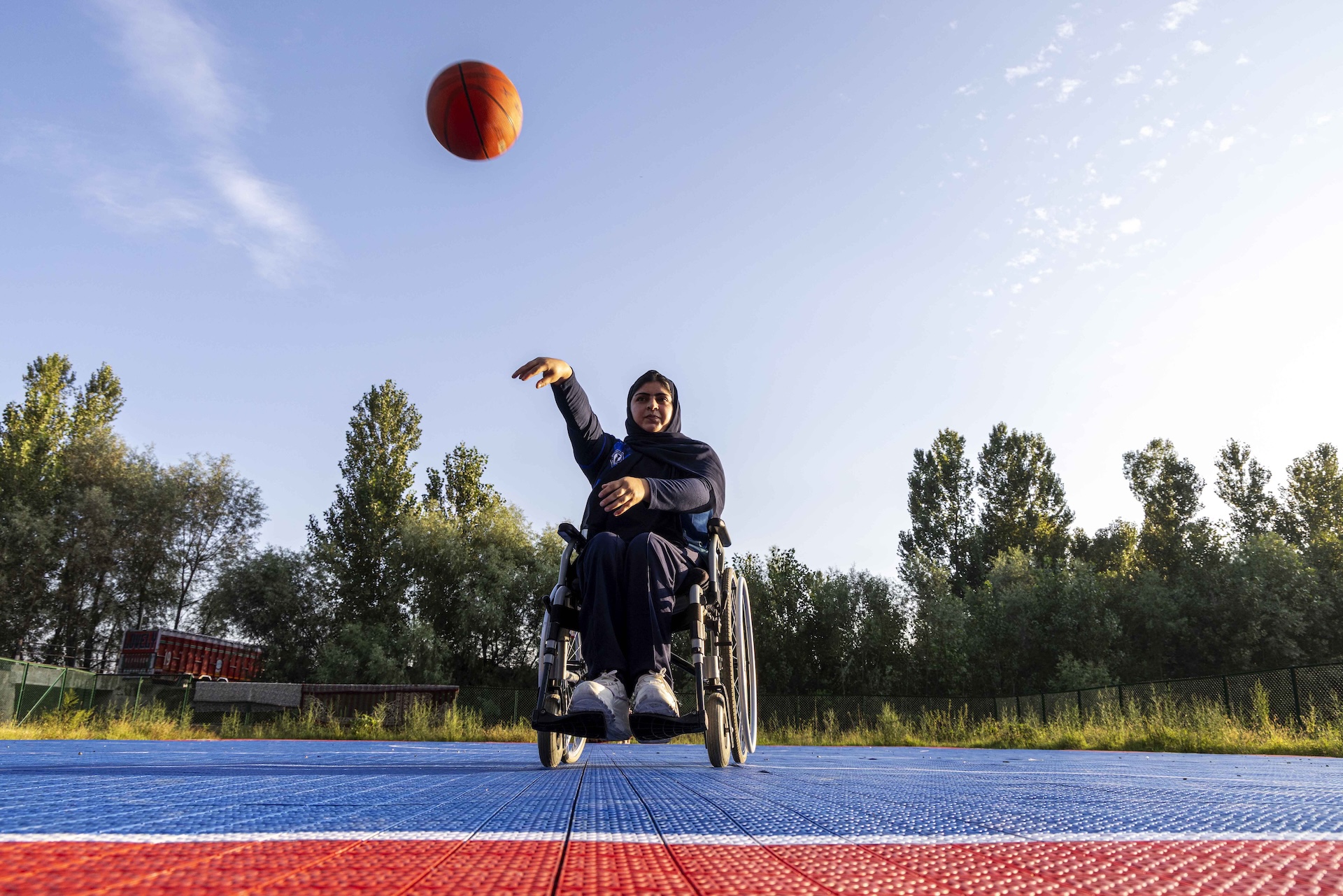 Faiqa Gulzar (27) sits in her wheelchair on an outdoor basketball court, facing the camera. Her right arm is extended and a red basketball is suspended in the air above and ahead of her. The sky is pale blue with a few fleecy clouds. The court is paved with sapphire blue tiles. In front of where Faiqa sits is the area beneath the hoop, which is paved with red tiles. Faiqa is dressed in black pants, a full-sleeved black top, a black hijab that covers her head and shoulders, and white sports shoes. There is a row of tall green trees in the far background, edging the perimeter of the court.