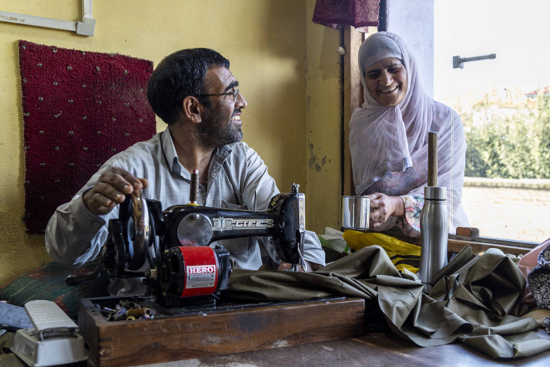Mid shot of Fayaz Ahmad Misger (46) who is sitting at his black table-top sewing machine while his wife Mumtaz Begum (40) stands facing him. Both are grinning at each other. Fayaz has turned his head sideways to look at Mumtaz who is on the other side of a large, open window. The sliding glass is half open and her upper body is visible as she leans on the window sill. Fayaz wears specs and a light grey kurta. He has cropped hair and a stubbly beard and moustache. Mumtaz has wrapped a pale lilac transparent dupatta around her head and shoulders, with a floral printed kameez showing through. She holds a steel mug in her left hand. The wall behind Fayaz is dark cream. There is a swathe of shiny chocolate-brown fabric beneath the sewing machine needle that spills across the faded brown wooden table.