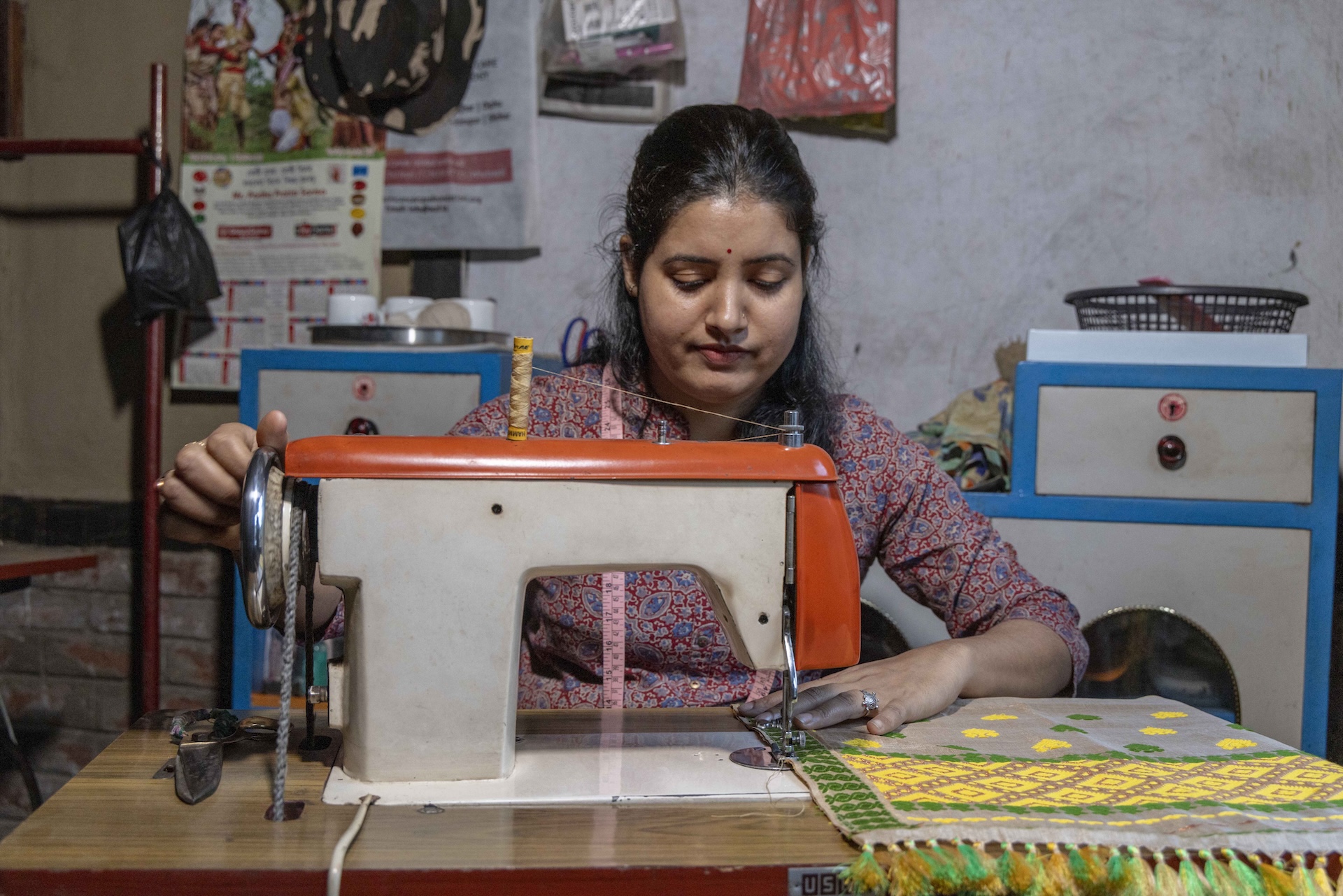 Gayatree (24) sits behind a sewing machine on a tabletop with a teakwood finish. Her black hair is backcombed and there is a tiny red bindi in the centre of her forehead. Her kameez has a Chinese collar, V neck and three-fourth sleeves. It is printed in a design of small floral sky-blue motifs of different sizes, on a red background. The machine is off-white with a clay-coloured top and side. Her right hand is on the wheel and her left hand next to the needle. She is stitching the side of a piece of light grey fabric with tassels at the edge. The cloth is embroidered in a geometric pattern of light green and dark cream.