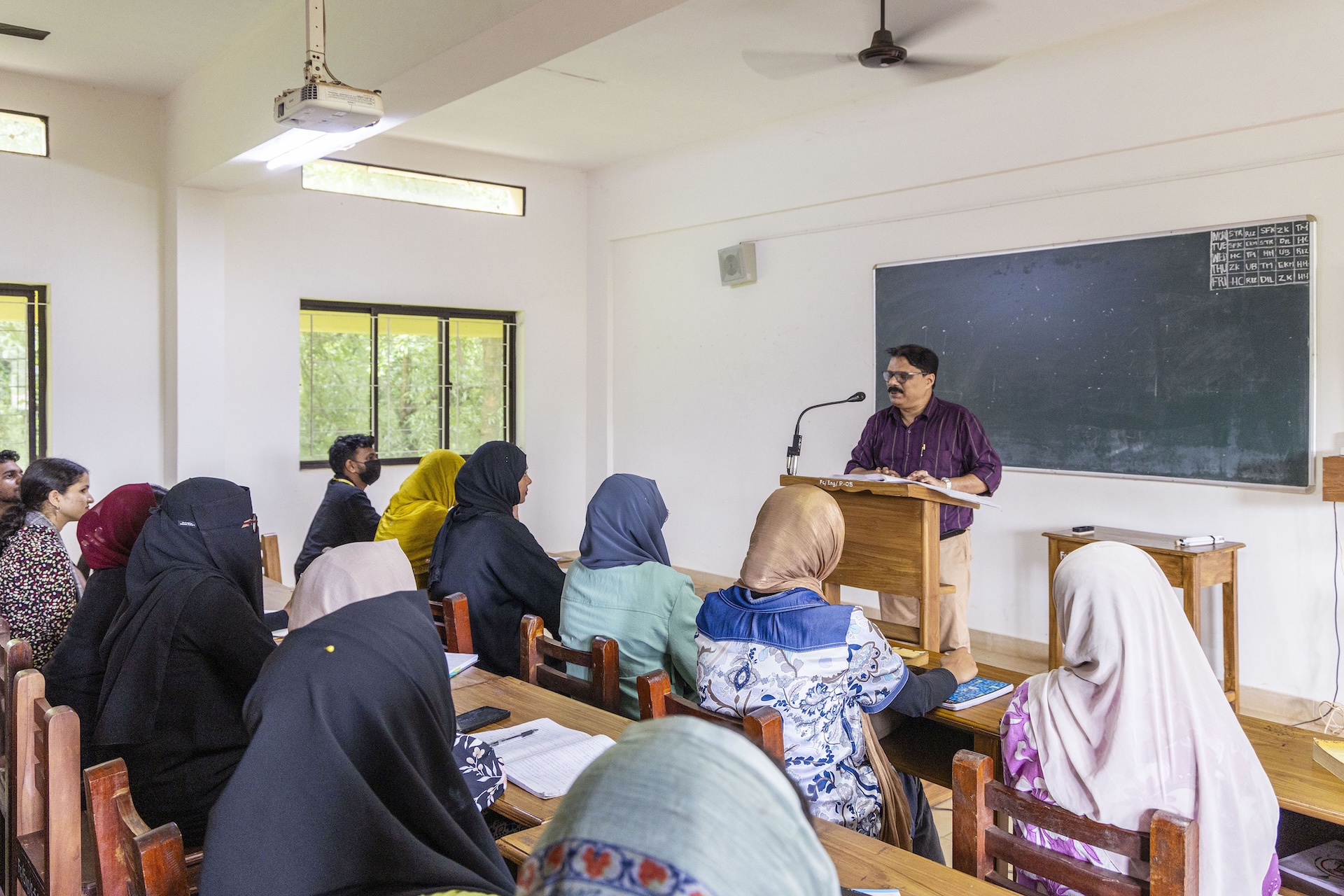 Habeeb C (43) stands behind a teakwood lectern in a classroom with off-white walls. His short, black, wavy hair has a centre parting, and he has a thick black moustache. His spectacles have a brown rectangular frame. He wears a deep purple shirt with thin beige stripes and sleeves rolled up to the elbow. The shirt is tucked into biscuit-coloured trousers. A large blackboard is fixed into the wall behind him. In front of him, students sit in teakwood chairs behind teakwood desks with their backs to the camera. The two rows in the frame have mostly young women. There is a young man sitting at the end of each row. Most of the women wear hijab in colours of beige, black, gold, red and dark grey. Three are dressed in black burka.