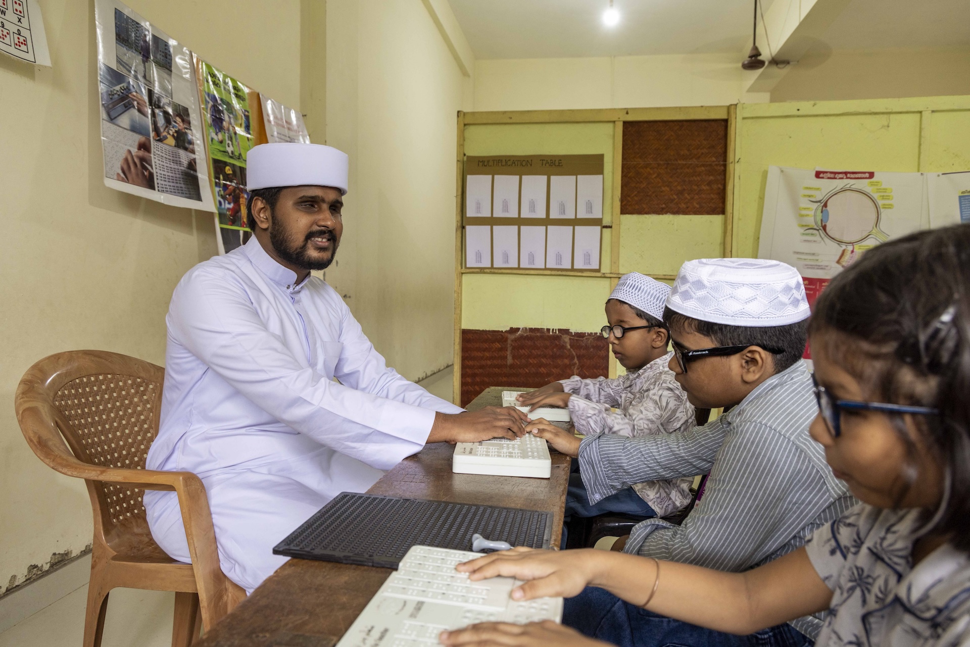 Side shot of Shabeer Ali Adany (25) sitting in a light brown plastic chair in front of a long teakwood desk in a classroom with cream walls. Three children sit behind the desk: two boys and a girl. Shabeer wears a white cotton kuppayam (a long robe) with full sleeves, a Chinese collar and buttons down the front. There is a white cotton circular cap on his head. He has a moustache that curves down and joins his narrow beard that lines his cheeks and chin. All three children wear specs with black frames. The boys wear white lacy prayer caps and full-sleeved jubbas. The girl has straight bobbed hair with a hair clip pinned to the side. Each has placed both hands on a white braille board in front of them. Shabeer has placed his hands on one of the boys’ braille board.