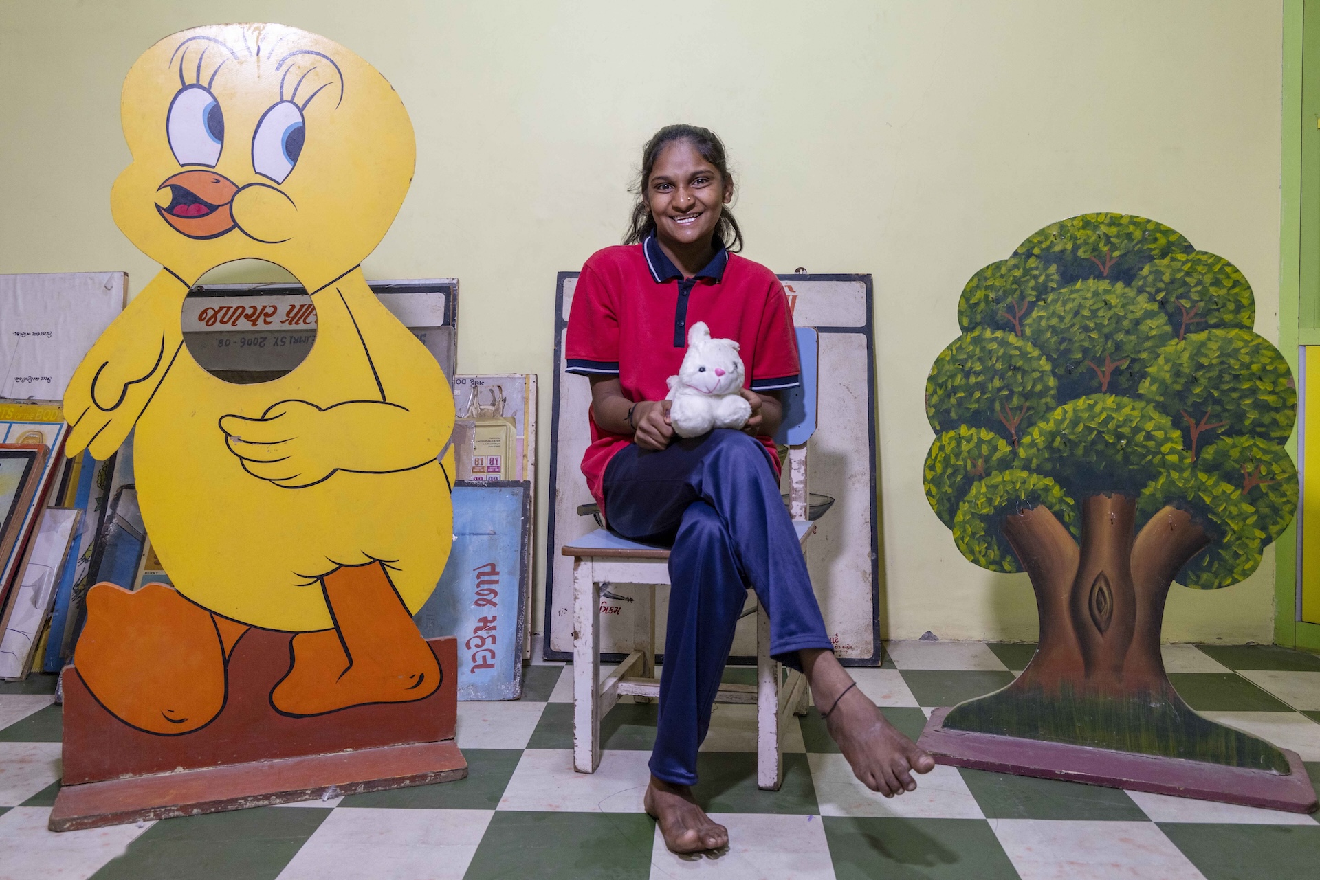 A smiling Hiral Kanaksinh (16) sits in a wooden chair painted white. Her long black hair is tied back in a ponytail. The wall behind her is cream and the floor has alternating squares of olive green and pale grey tiles. Hiral wears navy blue synthetic track pants and a blood-red, half-sleeved knitted shirt with black collar and sleeve edges. She holds a white stuffed bunny rabbit on her lap. To the right there is a large wooden cut-out of an artistic representation of a tree with a fat, mud-brown trunk and several bunches of green leaves. To the left there is a giant wooden cut-out of a yellow cartoon duckling with orange feet and beak and large oval eyes outlined in black with white eyeballs, black pupils, and black eyelashes.