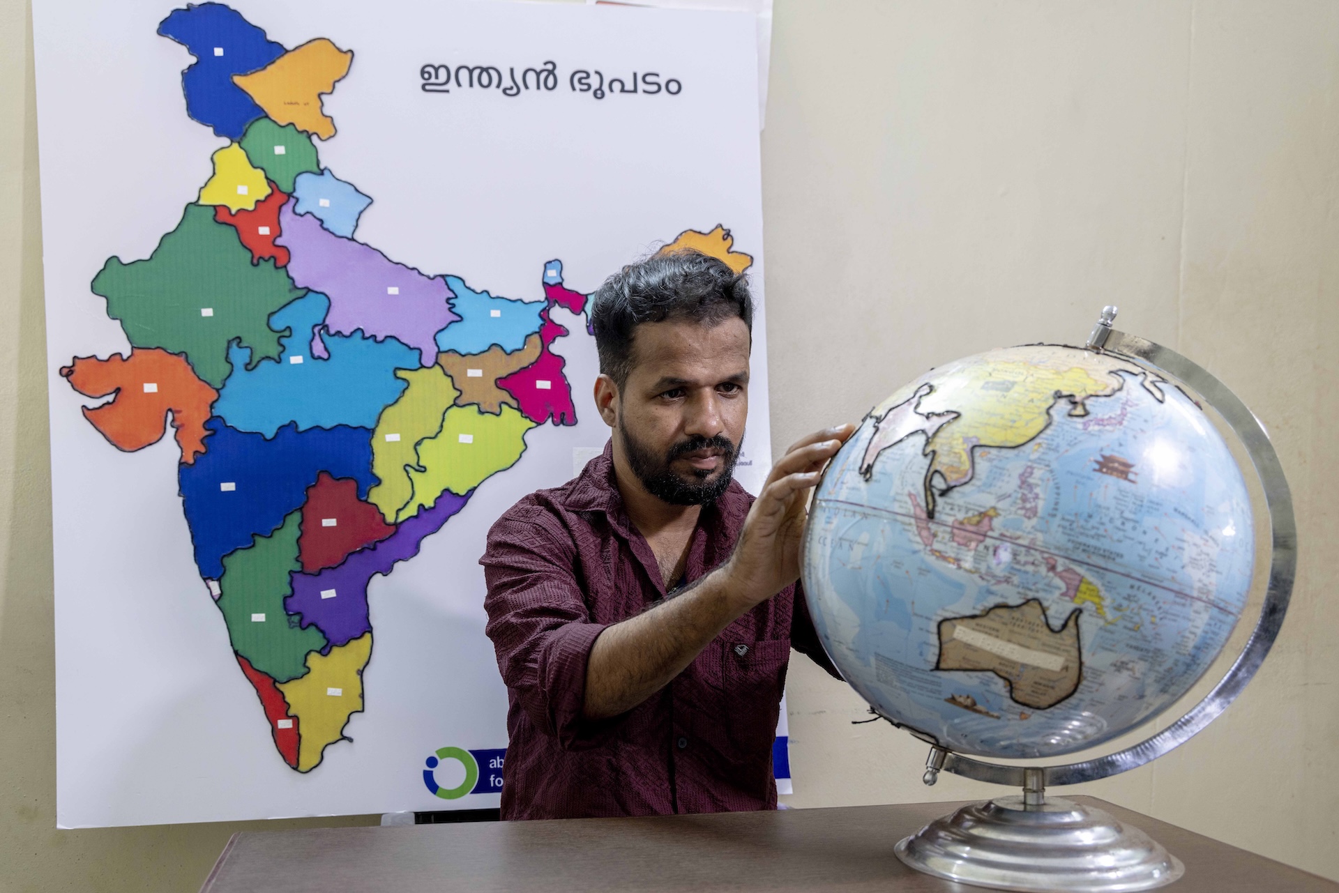 Mid shot of Jaisal A (30) sitting behind a light brown wooden table. There is an educational globe on the table. The continents have raised, black outlines. Jaisal is feeling the surface of the globe. He wears a maroon shirt with sleeves rolled up to the elbows.  He has straight, black, backcombed hair, a trimmed beard and a moustache. Behind him there is a large white chart with a multi-coloured map of India . The states have black outlines and are coloured sky blue, sapphire blue, leaf green, crimson, vermilion, yellow, orange, maroon, beige, violet, lilac, and light green. On the chart’s top right corner are large Malayalam letters that spell out ‘Indian bhoopadam (map)’.
