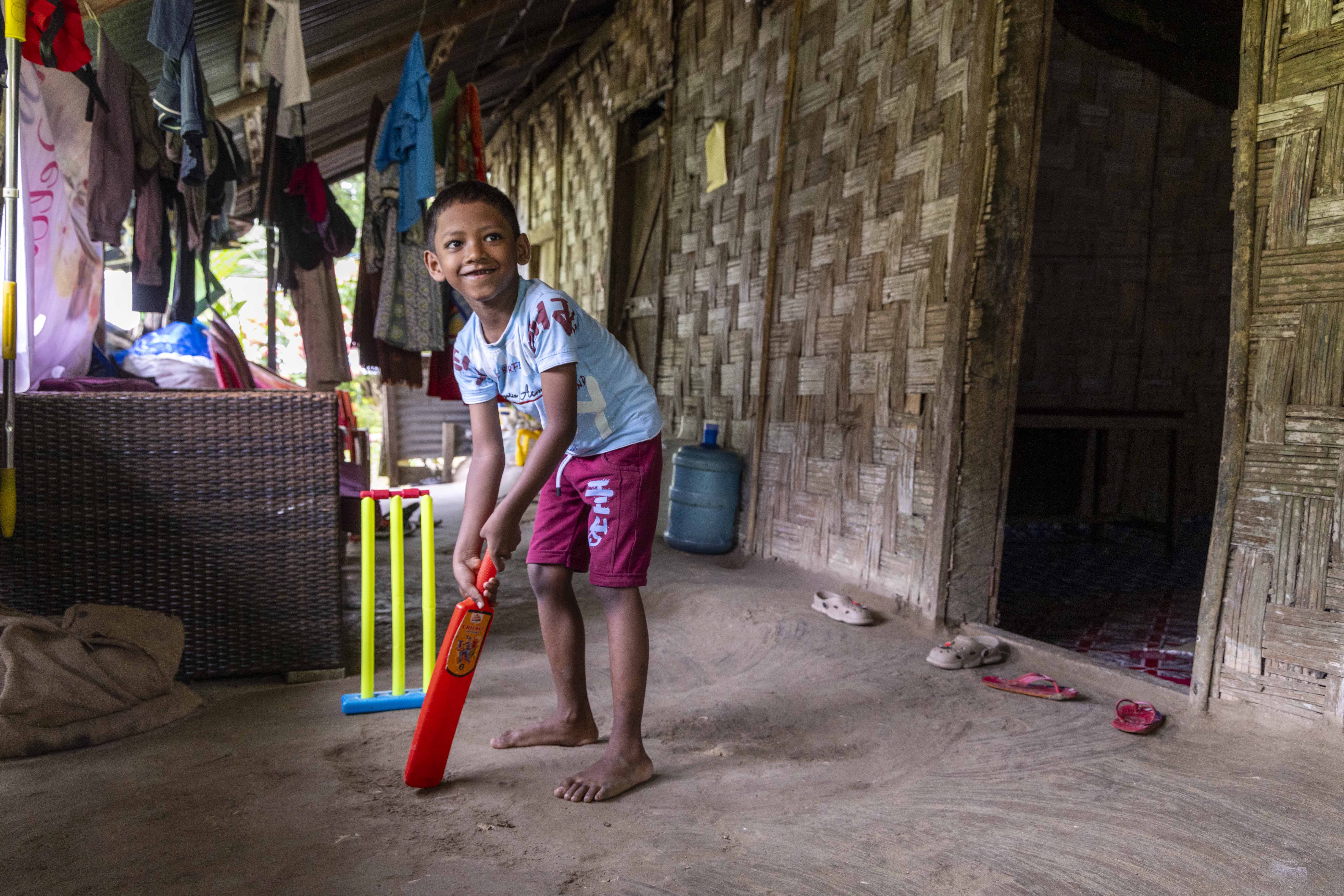 Joy Kerketta (8) stands in the veranda of his house, holding an orange plastic cricket bat and taking up a batting stance in front of three yellow plastic wicket stumps with a blue base and red bales. He wears a light sky blue T-shirt and maroon shorts and his hair is closely cropped. The mud floor of the veranda is beige and so is the wall made of woven bamboo cane.