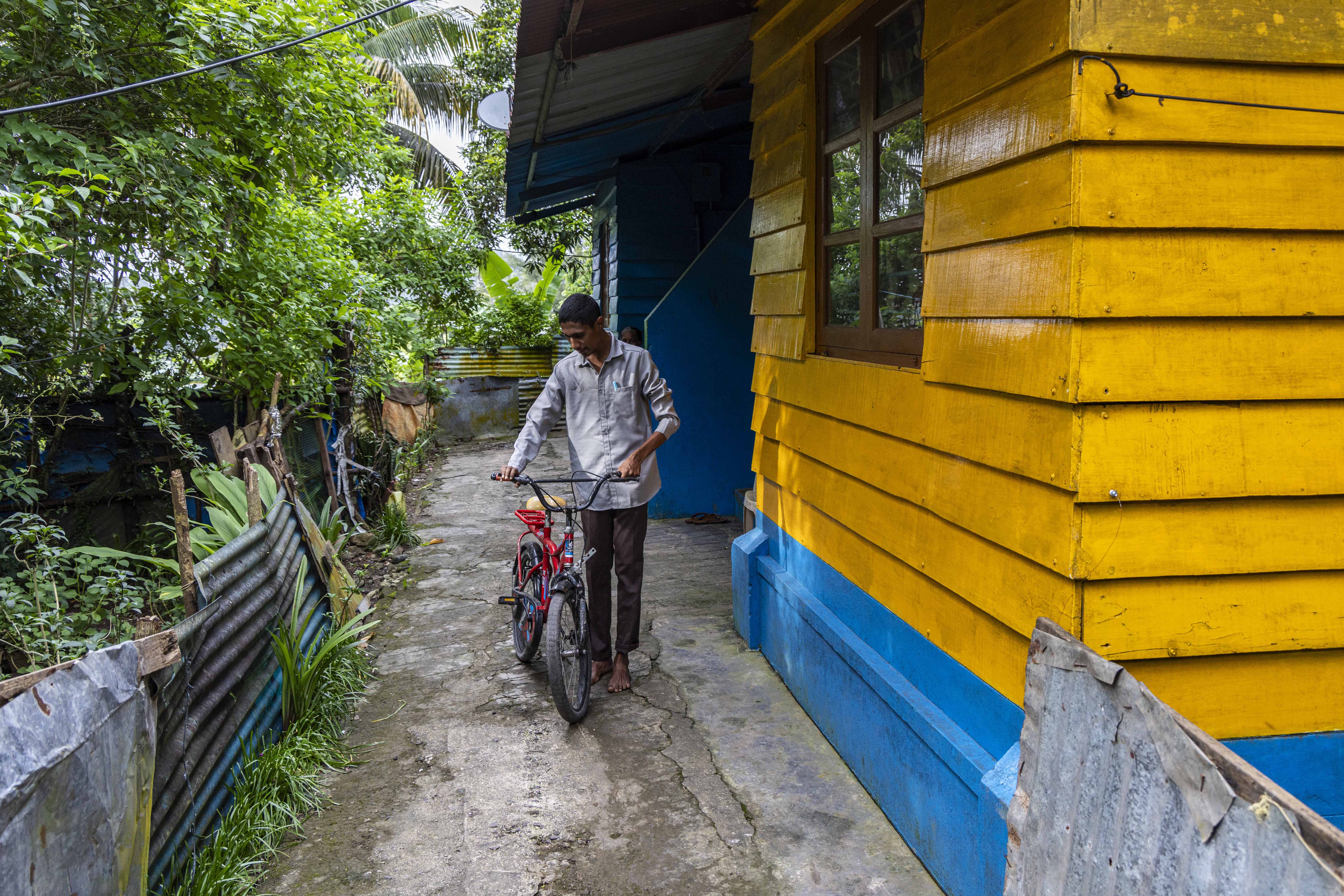 K. Parmesh (25) stands barefoot outside his house, looking down at his hands which grip the handlebars of a red, child-size cycle. His hair is cropped close to his head and he has a narrow, wispy moustache. He wears chocolate brown trousers and a full-sleeved smoky grey shirt. The façade of the house is made of slatted, horizontal wooden planks painted mustard yellow with a bright sky blue skirting. In the front wall there is a window with six square glass panes framed in wood painted chocolate brown. The uneven, light grey, narrow path on which he stands is roughly paved with cement. The low fence abutting the pathway is made of corrugated metal sheets. Behind the fence there is a row of green trees.