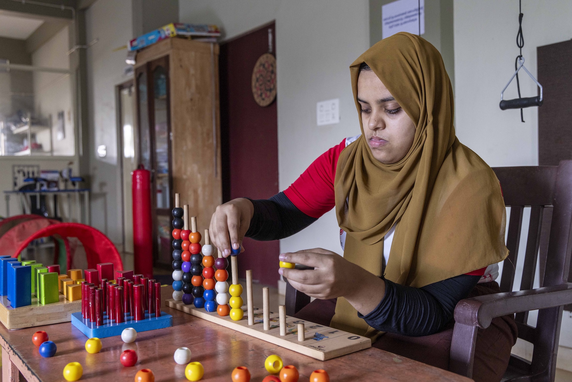 Mid shot of Lamiya Salim (24) sitting in a chocolate-brown chair in front of a mocha-brown rectangular wooden table. She wears a coffee-brown scarf that covers her head, shoulders and chest. The long sleeves of her top peep out from beneath the scarf. Half the sleeve, from shoulder to elbow, is blood red and the other half is black. There are pieces of motor-skill-building play equipment on the table. On a sky blue base there are red upright rods. There are also large orange, yellow and white beads scattered on the table. Just in front of Lamiya there is a plain beige wooden rectangular board fitted with a row of upright cigarette-shaped rods in ascending order of height. Five of the taller rods already have beads on them in random colours of orange, deep purple, violet, white, and dark sky blue. With her right hand Lamiya is slipping a blue bead onto a rod that already has three yellow beads on it. There is a yellow bead in her left hand.