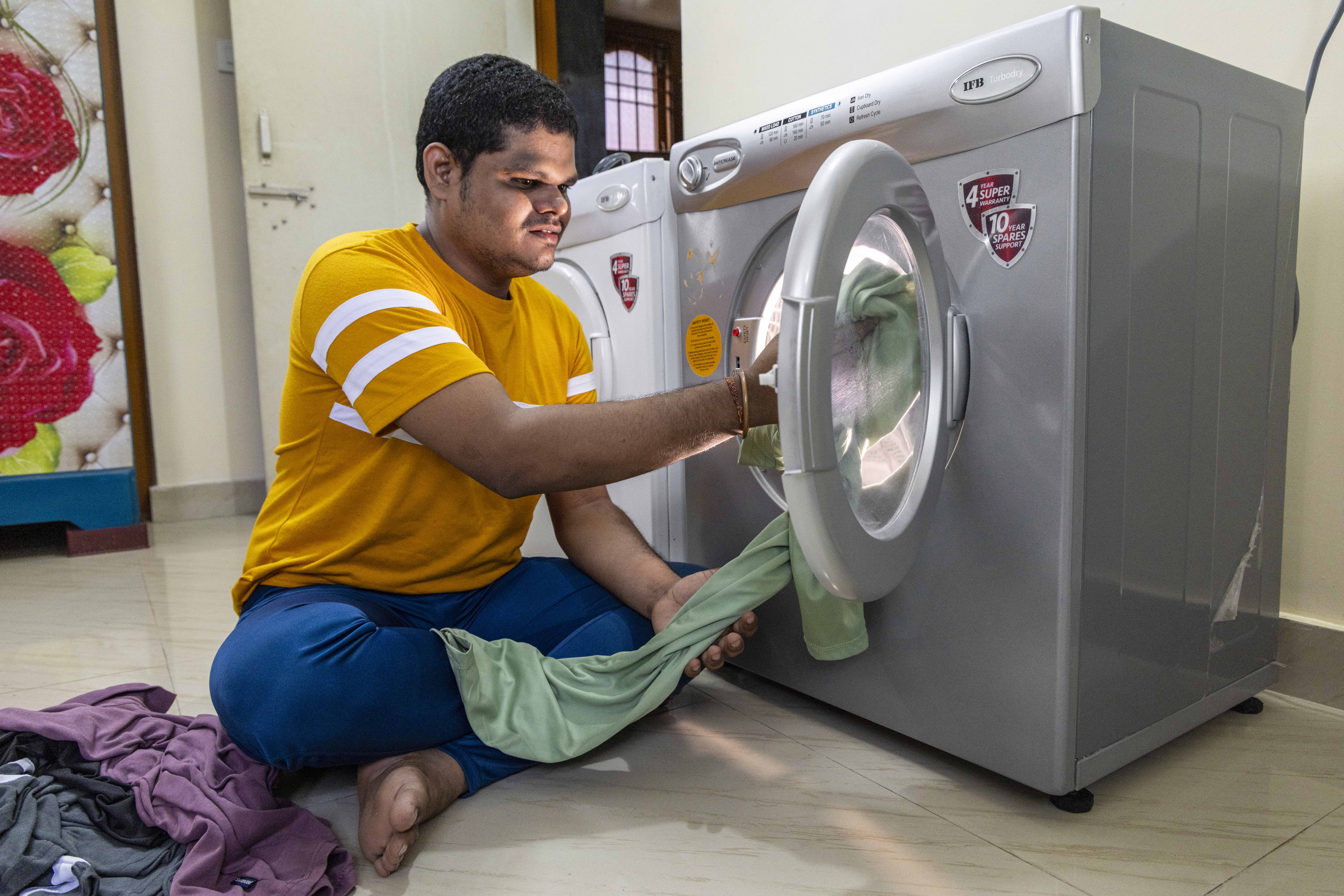 Laxmi Narayan (25), wearing a mustard T-shirt and sapphire-blue track pants, sits cross-legged on a white marble floor in front of a smoky-grey, front-loading washing machine. He is pushing a pista-green garment into the machine’s circular, open door. Next to his leg there are three crumpled garments coloured mauve, black, and ash-grey. His black, straight hair is cropped close to his head.