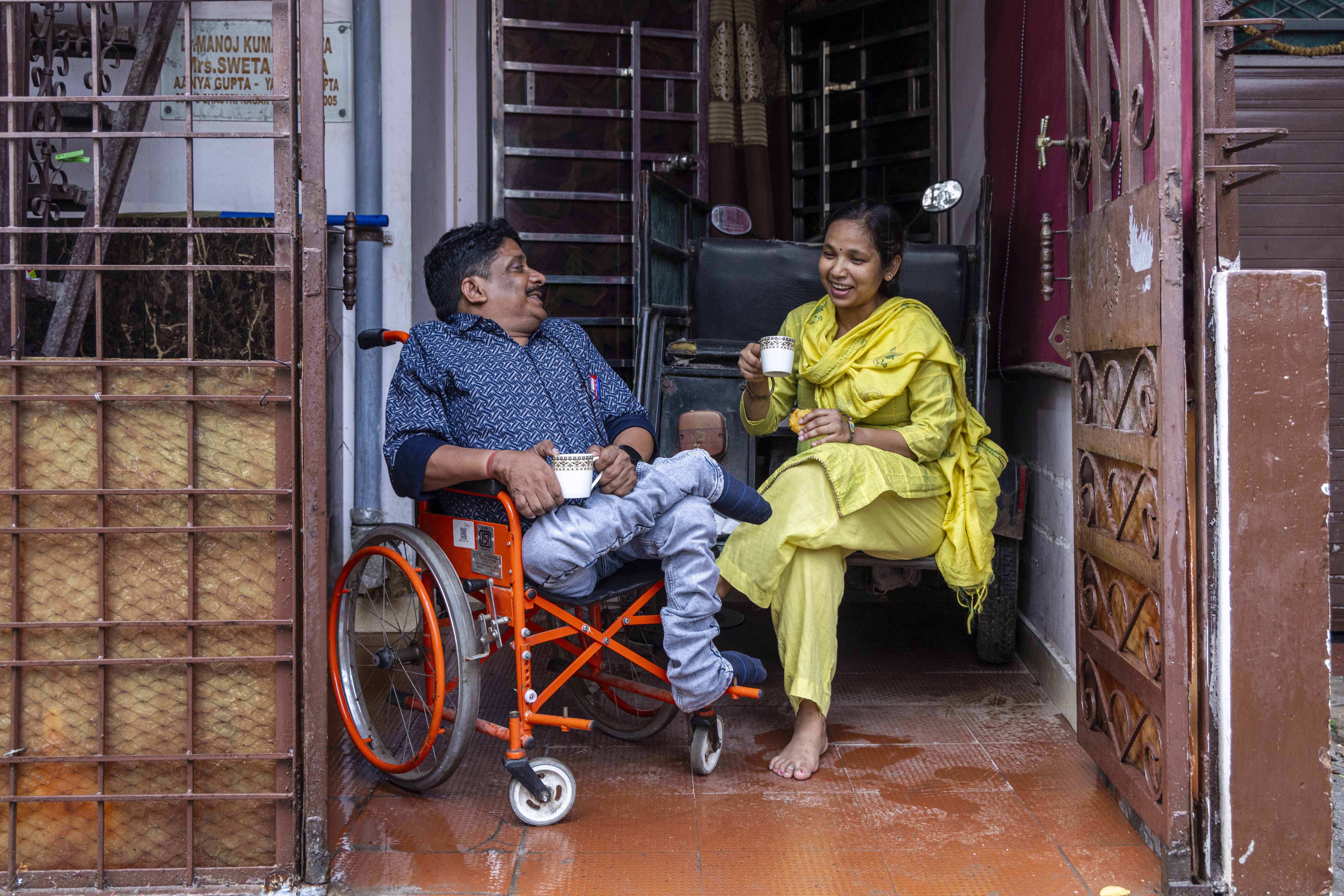 Manoj Kumar Gupta (50) and his wife Sweta Gupta (42) are looking at each other and smiling. Manoj sits in his wheelchair which has a metal frame painted vermilion and a black seat and backrest. Sweta sits in a chair upholstered in black leather. Both hold white porcelain cups; she holds the handle in her right hand and he holds it in his left while supporting the cup’s rim with his right. He has a thin black moustache and short black hair. He wears light blue jeans and a half-sleeved knitted shirt in a horizontal zigzag pattern of indigo and light blue with sleeves edged in broad navy blue bands. Sweta wears a light yellow salwar, matching dupatta, and a light yellow kameez printed with a very pale green pattern of squares filled with small dots. The floor is made of reddish-brown tiles.