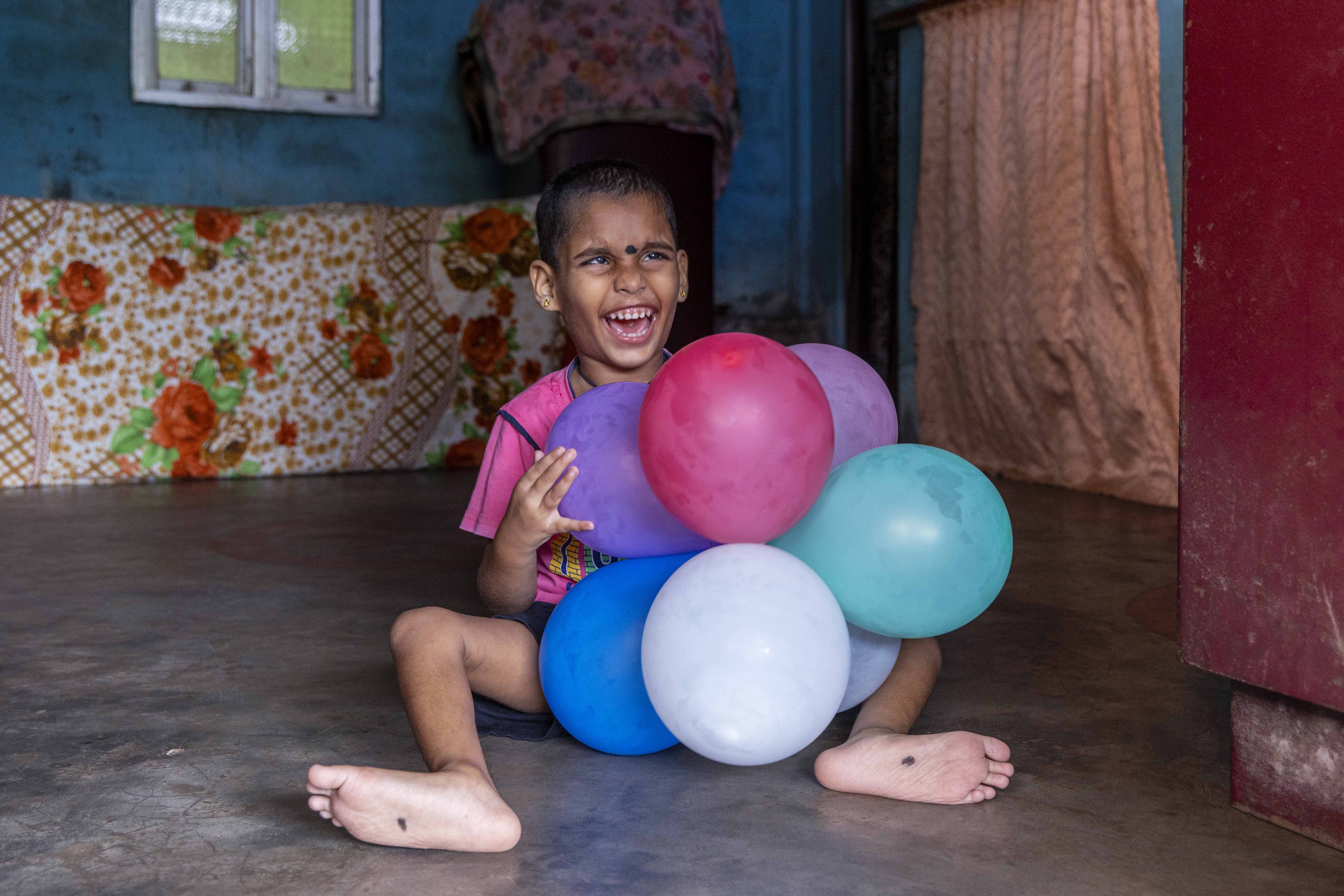 Manvi Kumari (6) sits on a polished, grey, plain cement floor with both legs bent at the knee and spread out, showing the soles of the feet. She wears a dark pink T-shirt and black shorts. Her hair is cropped close to the scalp. Her nose is crinkled and her mouth is wide open with teeth showing. A black ‘beauty spot’ between the eyebrows is meant to ward off the evil eye. The mid-sole of each foot is also marked with a black spot. She hugs a bunch of balloons tied together. They are red, green, white, blue, violet and grey.