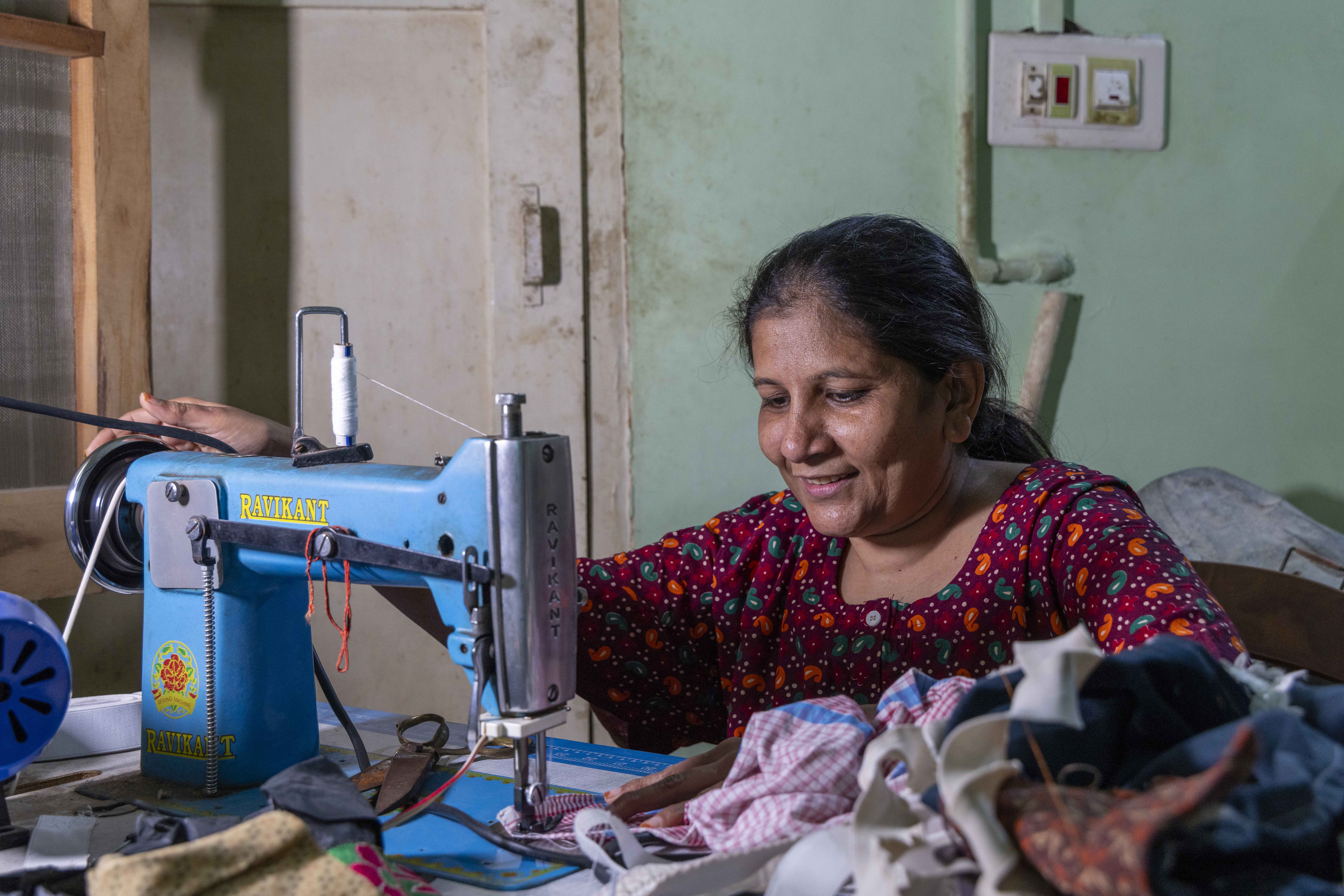 Mariyam Fakhruddin (46) sits behind a table on which there is a turquoise-coloured sewing machine with the brand name “RAVIKANT” stamped on the machine’s neck in yellow capitals. The wall behind her is a faded pista colour. Her black hair is backcombed and tied. She wears a half-sleeved round-necked maroon gown printed with small paisley motifs of sky blue and orange. Her right hand is on the wheel of the machine and her left hand holds a piece of fabric in place below the needle. An untidy heap of pieces of different coloured fabric is on the table next to her left hand.