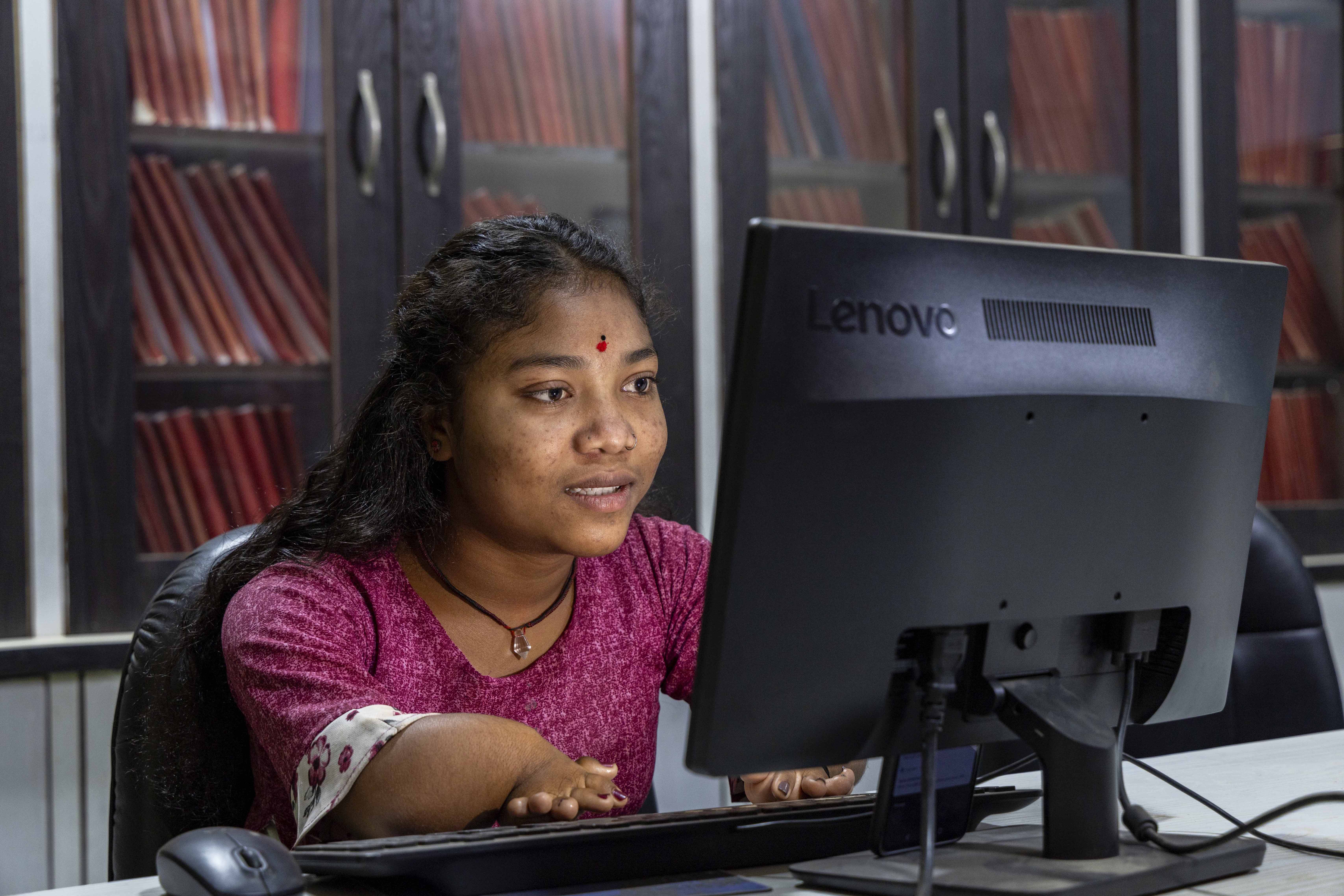 Mase Markam (15) sits in a black leather chair in front of a light grey desk, looking at the screen of a black desktop computer. Her hands, which are foreshortened, rest on the black keyboard. Her long curly hair with a centre parting flows down her back. On her forehead there is a small red bindi with a tiny black dot just above it. Her chain consists of a brown strand around her neck and hanging from it is a small, conical, transparent pendant. She wears a maroon round-necked top with half sleeves edged with a broad white frill printed with small maroon flowers. In a glass-paned brown wooden cupboard behind here there are three shelves stacked end to end with rows of folders with their red and maroon spines showing.