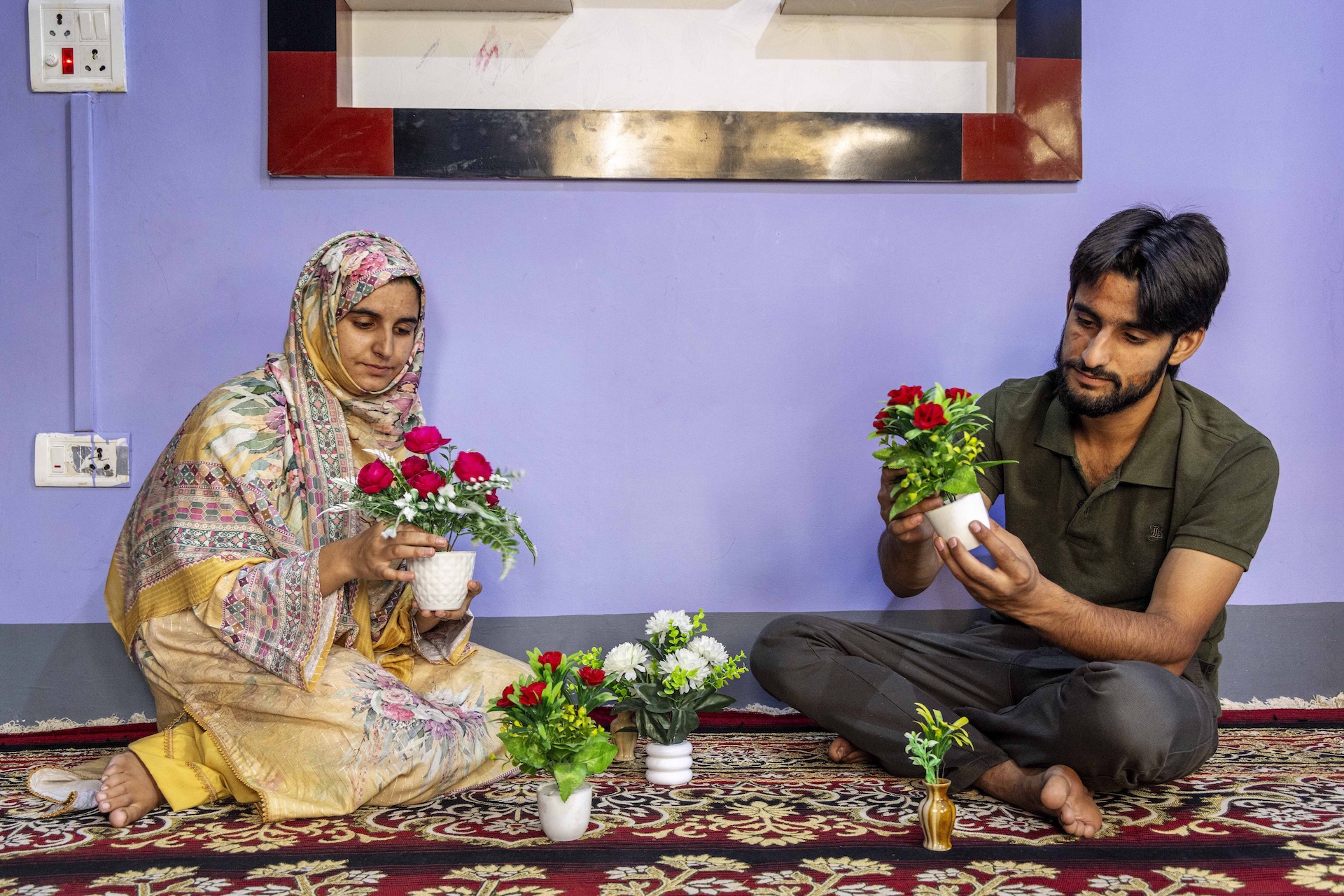 Ifla Syed (21) and her brother Mehran Syed (20) sit on a red carpet in a room with a lilac wall behind them. Ifla wears a sandalwood coloured salwar-kameez with a floral bouquet printed on the front of the kameez in pale pastels of pink and green. Her head scarf wrapped around her shoulders is also sandal-coloured printed with broad, ornate borders of pale pastel pink and green. Mehran has thick straight hair with a centre parting, a moustache, and a narrow beard that lines his face. He wears a dark olive-green half-sleeved knitted shirt and dark grey pants. The siblings each hold a small white pot of artificial crimson roses with green leaves. On the carpet in front of them there is a similar pot with similar flowers, a tiny brown vase with two artificial green stems and leaves, and a white pot shaped like three rings one on top of the other, containing white artificial chrysanthemums and green leaves.