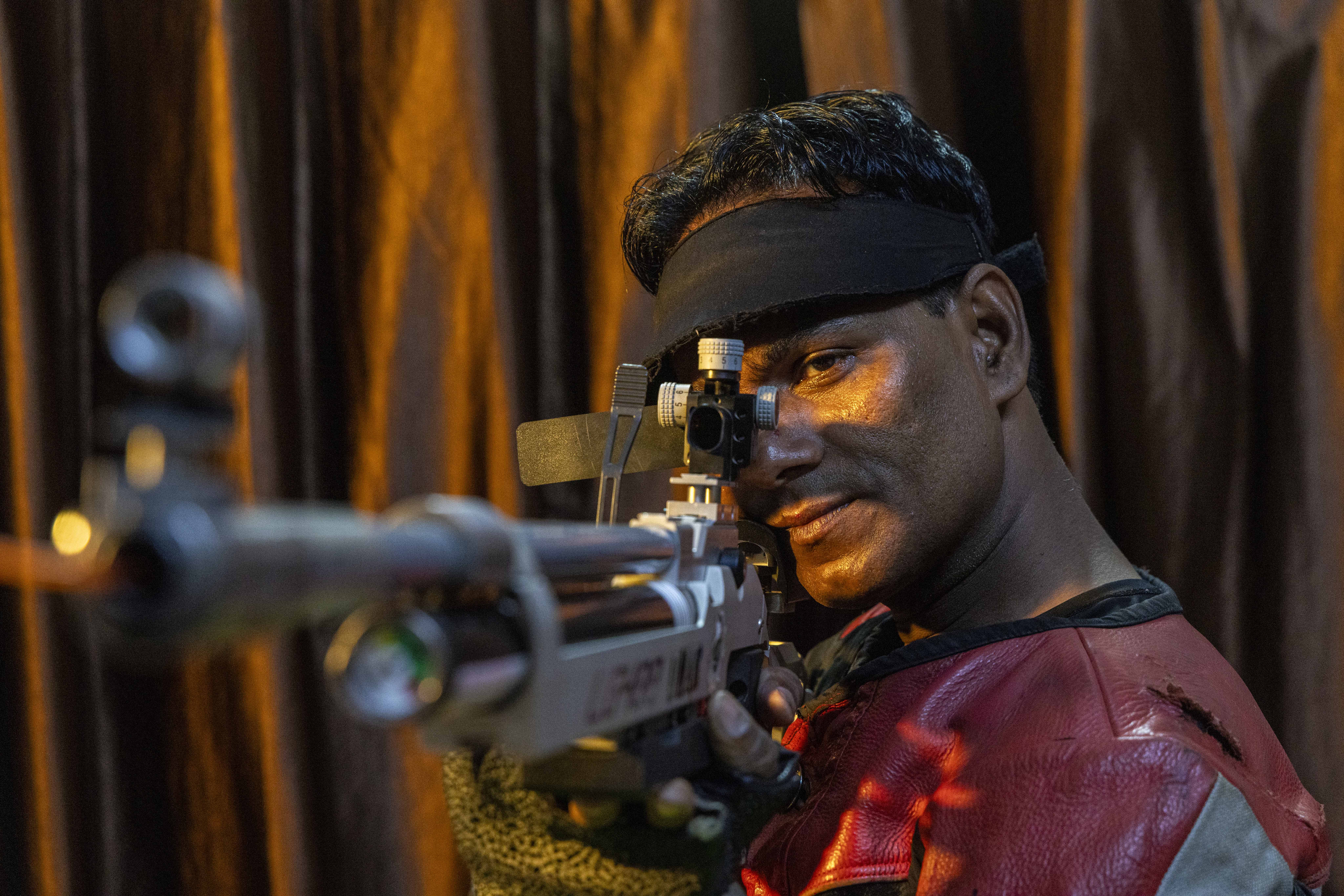 Mid shot of Mohamad Umar (38) aiming an air rifle at the camera. The barrel is tilted slightly to the left of the frame. Umar has short, curling black hair and is clean-shaven. He wears a red leather jacket. A black band encircles his forehead. His right eye is obscured by the rear sight of the rifle. His left eye is open and his right hand grips the trigger.