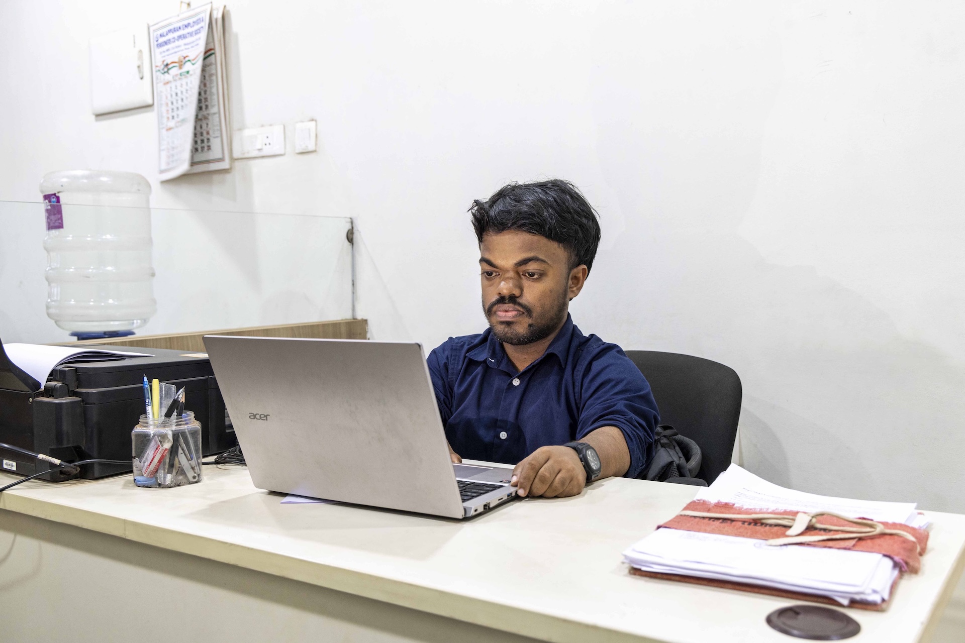Mohammed Raees (27) , wearing a bluish-black shirt with sleeves rolled up to the elbows, sits in a dark chocolate computer chair behind a long white desk. The wall behind him is ivory. He has a narrow black moustache that curves around his lips and a thin, narrow, black beard that covers his chin and cheeks. There is a silver laptop open in front of him and his hands are on the black keyboard. 