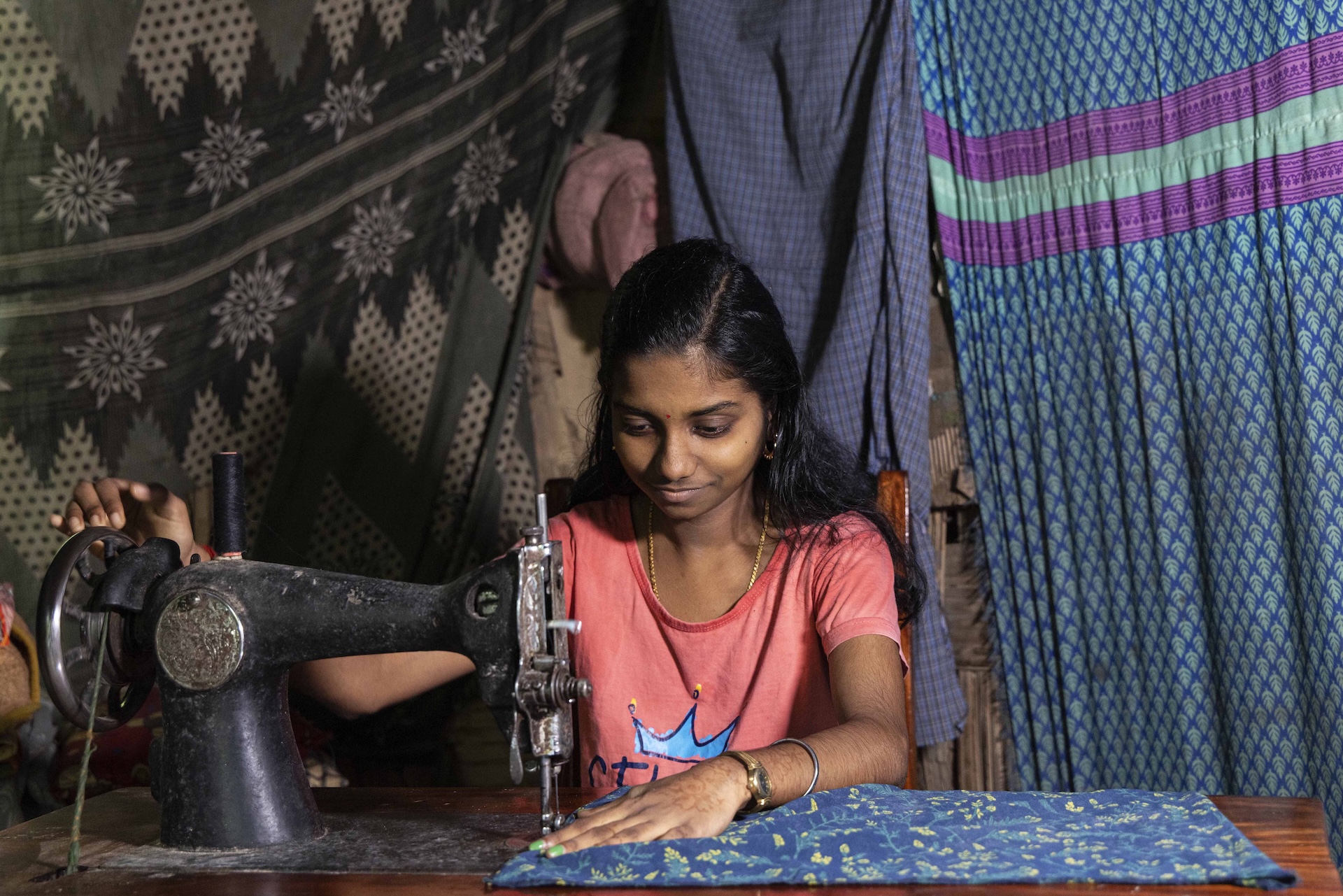 Mid shot of N. Deepika (18) sitting behind an old-fashioned, black, foot-operated, treadle sewing machine with a tan-coloured wooden table. She wears a rose-pink T-shirt and her long black hair has a side parting and flows down her back and shoulders. Her right hand is on the wheel and her left hand holds a piece of dark sky blue fabric in place beneath the needle. 