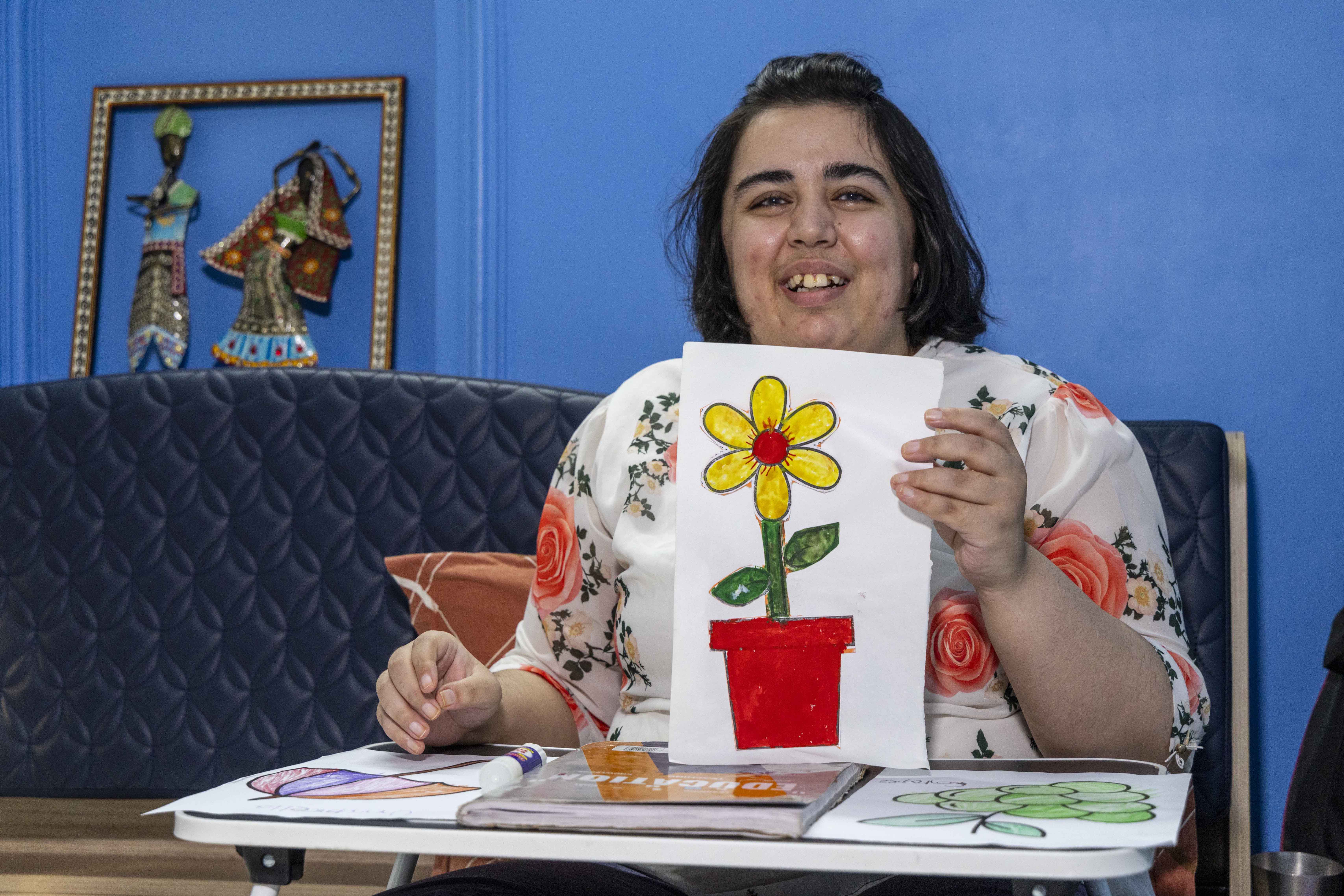 Mid shot of Nikita Gulati (29) sitting behind a small white folding table, smiling broadly and holding up a drawing on a white sheet of paper. She has bobbed hair with a section of the centre combed back and the hair on either side falling over her ears. She wears an off-white synthetic top printed with large orange roses and tiny grey leaves. The drawing shows a red flower pot that holds a single flower. At the top of a thick, dark green stem with two green leaves, one on either side, there is a yellow flower with six oval petals and a red centre. The sofa she sits on has a black quilted leather backrest and the wall behind it is a bright sky blue.