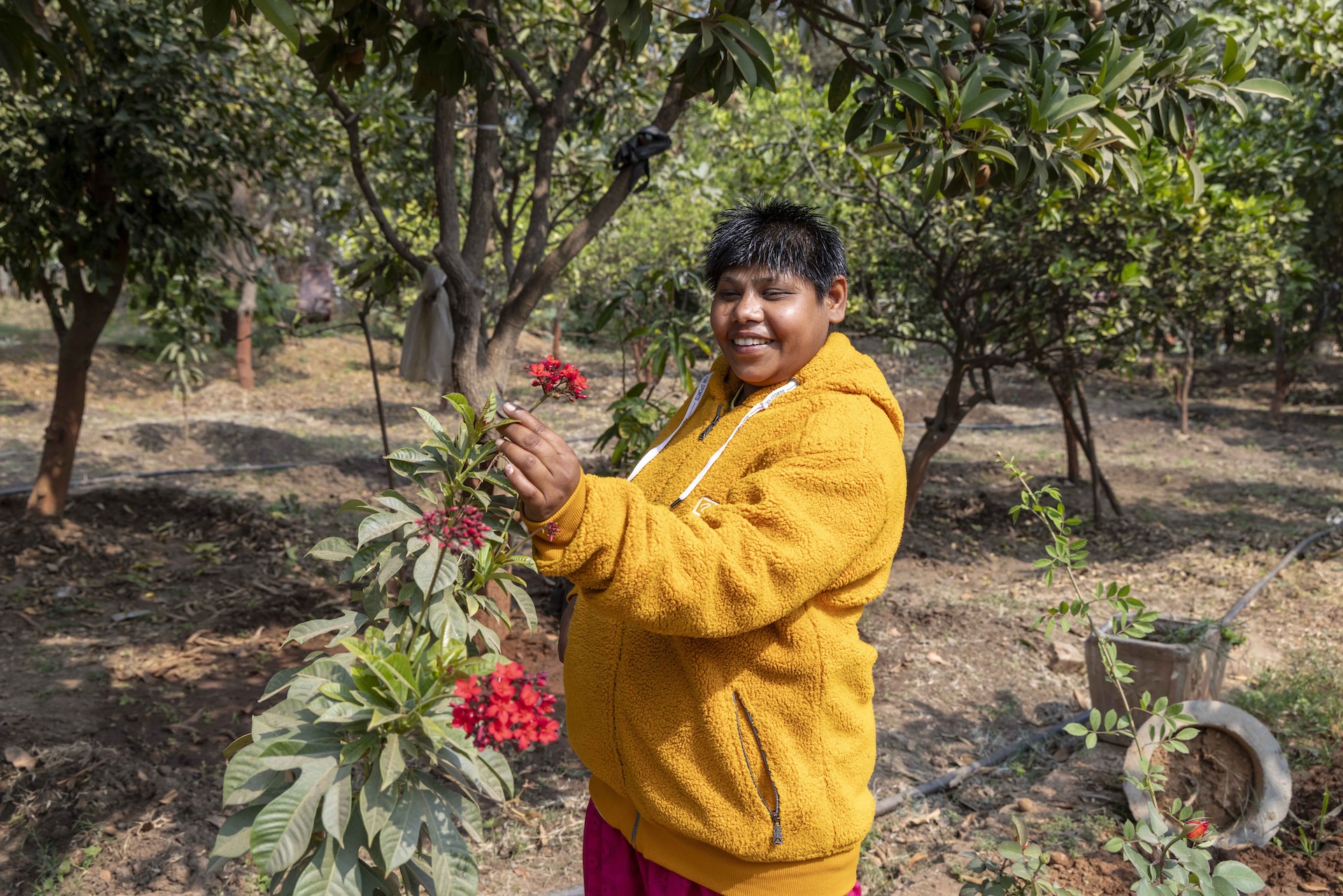 Pintuben Meda (19) stands in a shady grove, in the midst of guava trees, next to a bush with red inflorescence. She wears crimson pants and a mustard-coloured chunky cotton cardigan with zippered side-pockets, a zip-up front, and a hoodie. The hoodie is pushed back, revealing black spiky hair. She smiles as she holds the stem of a spikelet of red flowers.