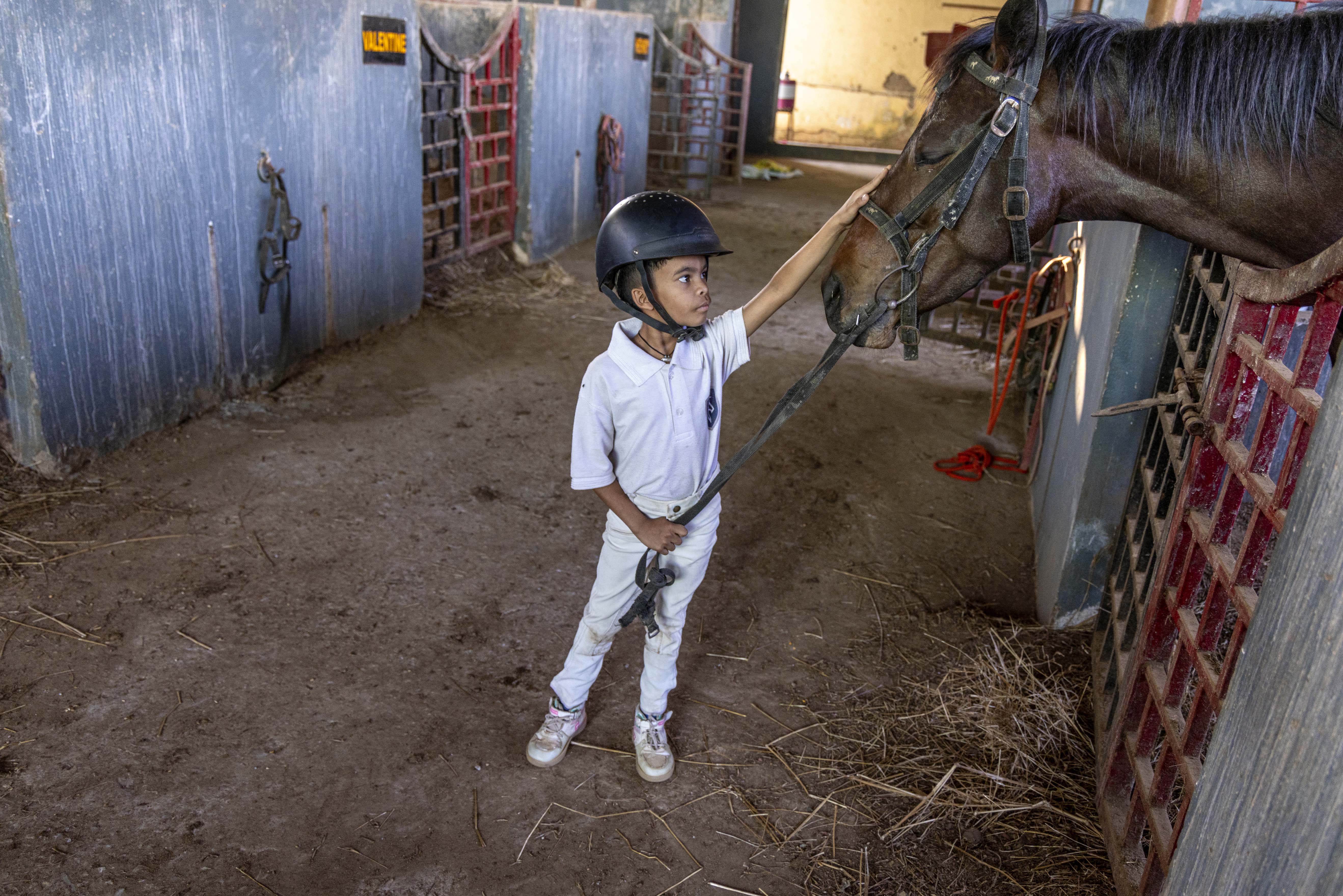 Prince Yalam (11) stands in a stable, petting the head of a chocolate brown horse leaning out of its stall. Bits of straw are strewn around the muddy floor. Prince wears a black riding helmet with chin straps, a white half-sleeved knitted shirt with collar, tight white trousers, and white shoes. Pulling forward the horse’s reins with his right hand, he is stroking its muzzle with his left.