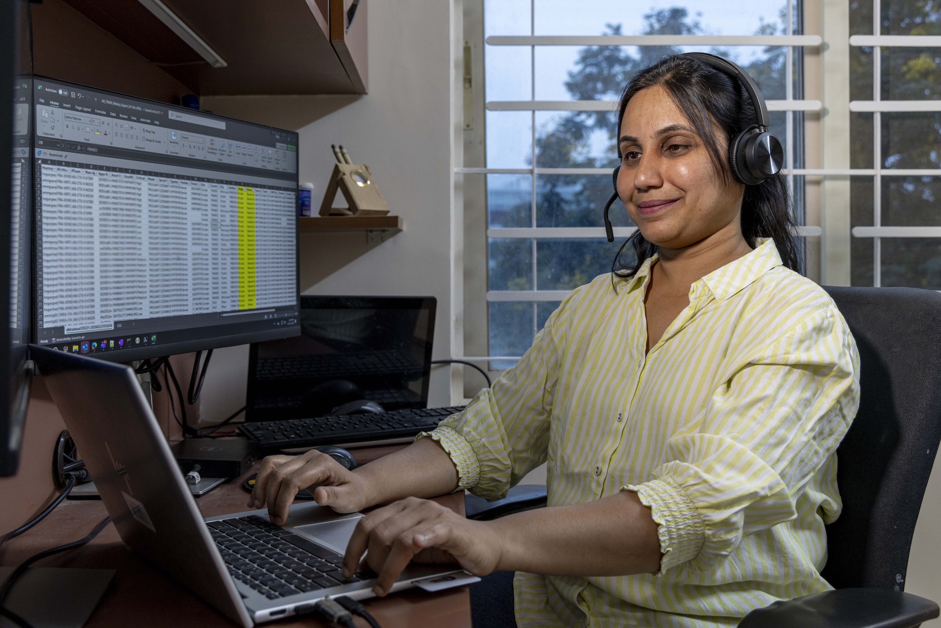 Mid shot of Priyanka Agarwal (42) sitting in a black computer-chair in front of a silver-coloured laptop. The laptop is on a brown wooden table and her fingers are on the black keyboard. To the right of the laptop there is a large computer monitor with text showing on the screen. Priyanka’s long, straight black hair is parted in the middle and she wears a pair of black headphones. Her top has thin horizontal stripes of pale yellow and white and the sleeves, pushed up to the elbows, have frilly edges. The open window in the beige wall behind her has white horizontal bars.