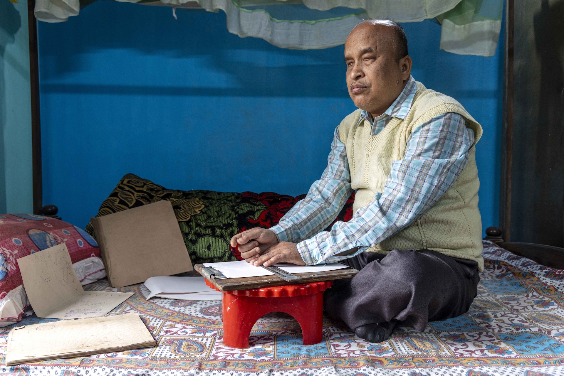 Pradeep Dowarah (52) sits cross-legged on a bed with a red plastic footstool in front of him. He is bald with close-cropped hair ringing the back of his head. He has a pencil-thin moustache and his eyes are almost closed. He wears dark grey trousers and a sleeveless ivory-white sweater over a full-sleeved shirt in a plaid pattern of sky-blue and light grey checks. The wall behind the bed is dark sky blue. The bedsheet is printed in a swirling pattern of sky blue, white, and pale pink. There is a thick, rectangular, brown wooden board on the stool. The head of the board is fitted with a broad steel clip. There is a foolscap sheet clipped to the board and Prodeep has placed both hands on a tactile braille scale placed horizontally in the middle of the sheet. There are several white braille sheets on the bed.