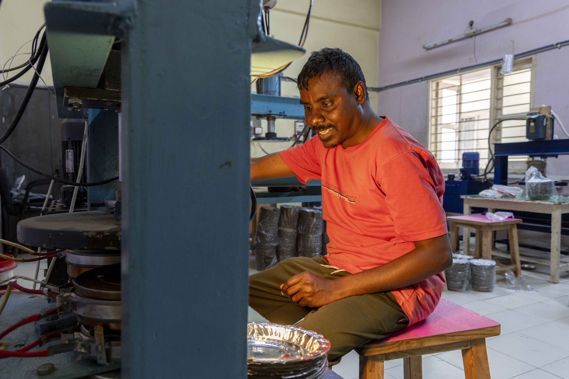 Side shot of Rajesh Maganbhai (28), wearing an orange T-shirt and dark brown trousers, sitting in front of a paper-plate-making machine. He has a bristly moustache and a sparse, bristly beard on his chin. One upright, broad, grey-painted, rectangular iron bar of the nether side of the machine is visible, but the rest is hidden from view. Next to the lower end of this bar there is a stack of white paper plates with raised rims. The plates are lined with silver foil. In the background there are several stacks of paper plates, a couple of plain wooden tables, and a plate-making machine with a grey horizontal iron bar resting on two vertical bars.