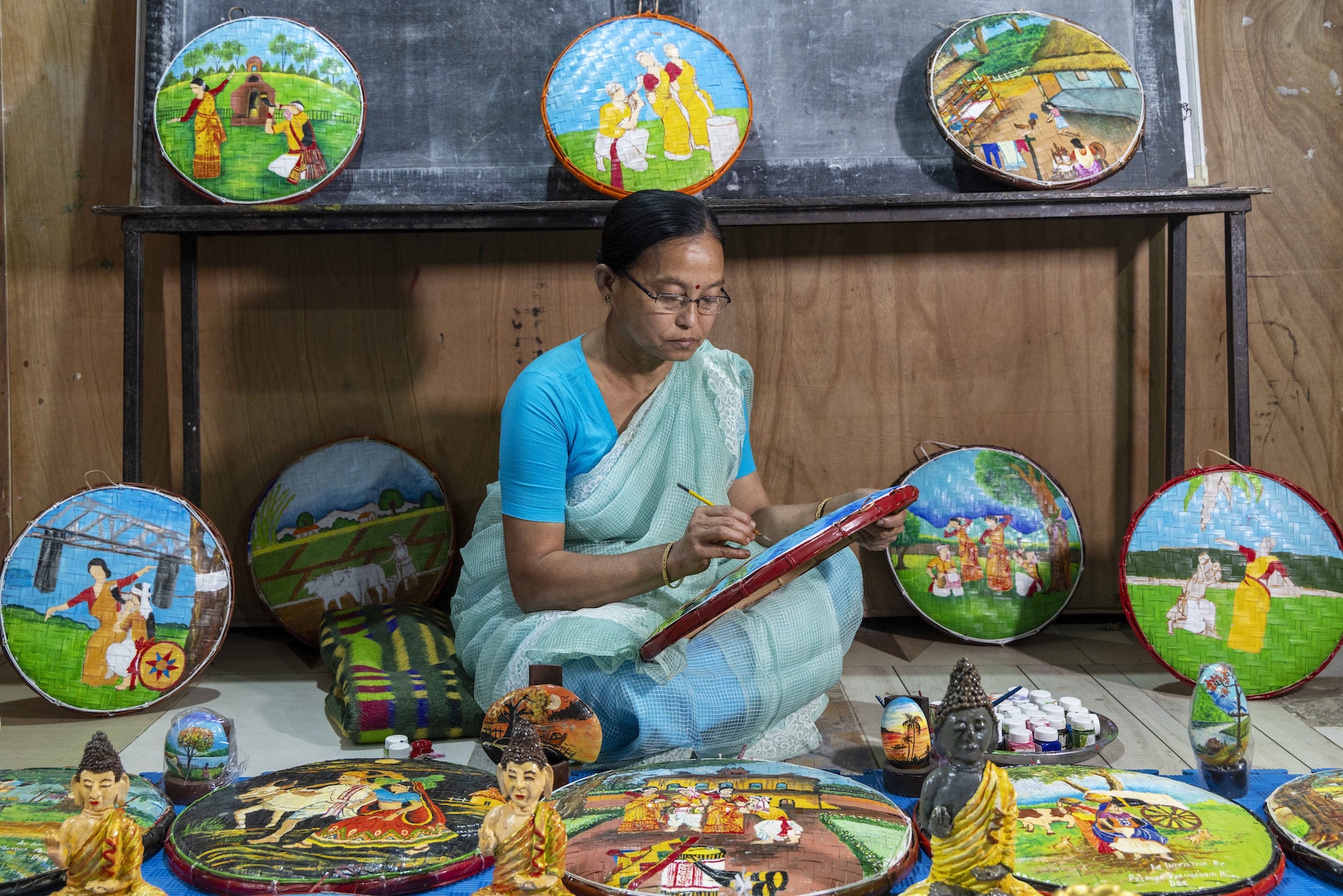 Rijumoni Chetia Gogoi (57) sits cross-legged on the floor. She wears a turquoise blue cotton blouse and a pale sky blue, semi-transparent, voile sari. Her black hair with a slightly off-centre parting is tightly combed back. She wears rectangular rimless specs and a small red bindi in the centre of her eyebrows. In her left hand she holds a disc made of woven cane which rests on her knees. The circular frame of the disc is painted maroon. Holding a thin yellow-handled artists’ paintbrush in her right hand she is applying paint on to the disc’s surface. There are two finished paintings on either side of her, on the floor, propped up against the coffee brown wall. Three more such paintings rest side by side against the wall at a height, on top of a narrow steel shelf painted dark grey. Placed flat on the floor side by side in front of her are three such paintings. In front of these paintings there are three small statuettes of the Buddha. All the paintings depict dancers in traditional Assamese attire dancing the Bihu on green grass against a blue sky.