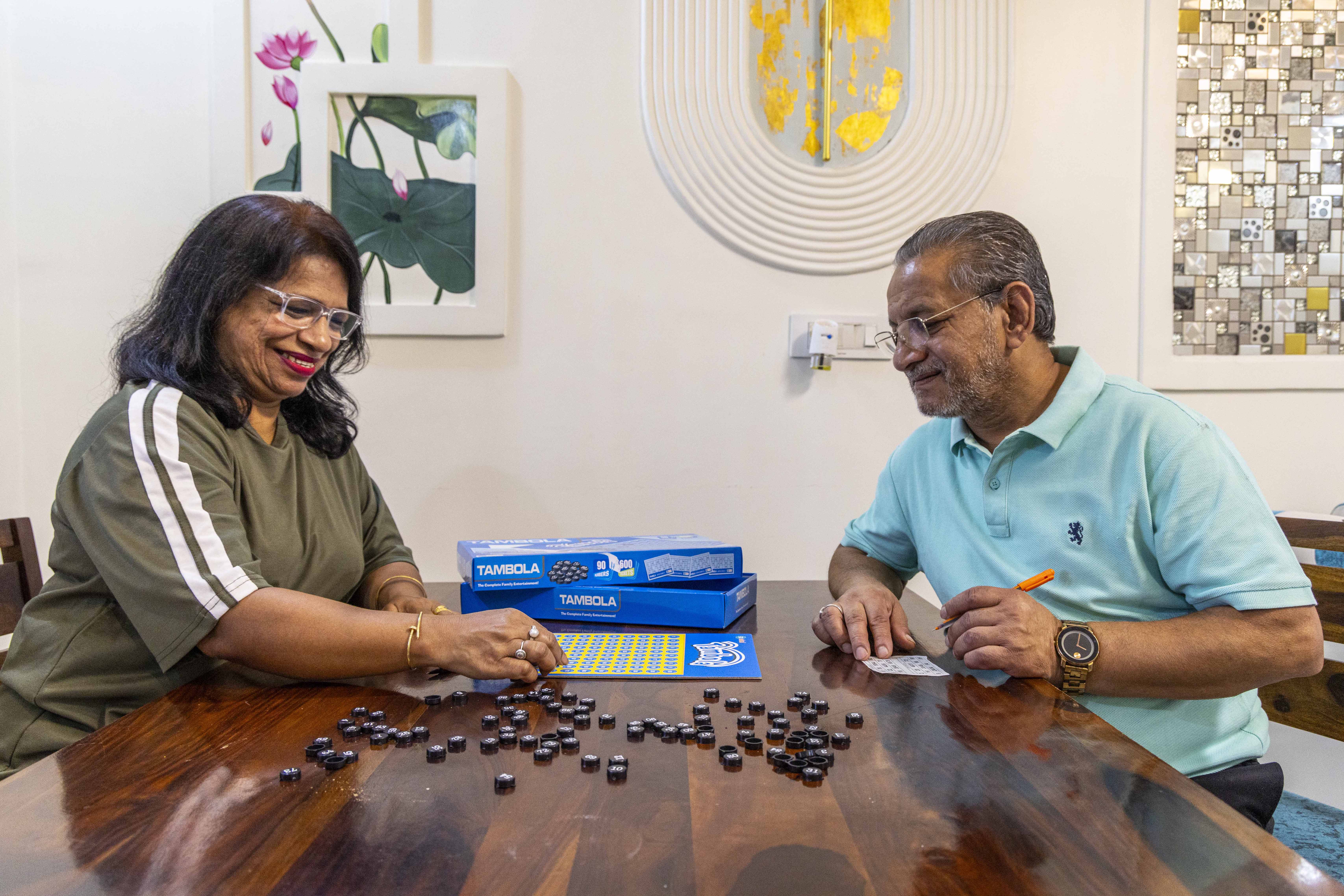 Sitting on opposite sides of a wooden table with a polished dark brown surface are Sangeeta Sharma (58) to the left and her husband Rakesh Sharma (61) to the right. There is an ivory wall behind them and a board game between them on the table. The board game is made of a thin flat square cardboard printed with small yellow squares on a sapphire blue background. Sapphire blue cardboard boxes with “TAMBOLA” in white capitals lie next to the board. Sangeeta wears an olive-green T-shirt with two broad white bands running down the sleeve. She has shoulder-length hair and wears crimson lipstick and specs with white frames. She is smiling broadly as she places a disc-shaped token on the board. There are several small brown discs with numbers on them scattered on the table next to the board. Rakesh wears a sky blue half-sleeved knitted shirt and specs with a thin metal frame. He has white stubble on his cheeks and above his upper lip. On a small white rectangular piece of paper he is keeping score with an orange pen held in his left hand.