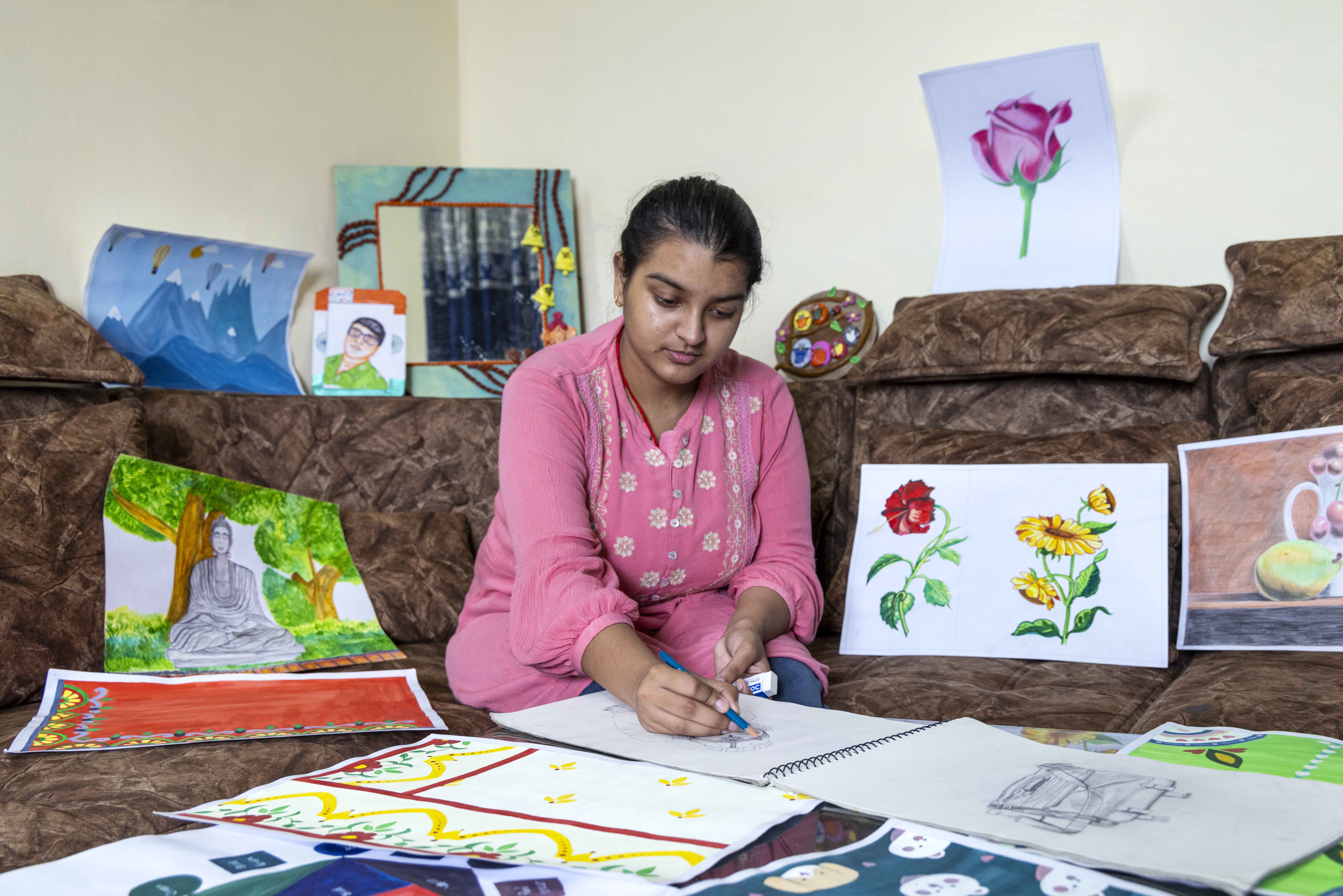 Raktima (19) sits on a sofa behind a large side-table. The sofa is upholstered in a chocolate brown fabric and the wall behind it is ivory-coloured. Raktima wears ash-grey pants and a rose-pink three-fourth-sleeved thigh-length top with white floral embroidery on the front. She is sketching with a black pencil on the white page of a large spiral-bound drawing pad in front of her. She is surrounded by her own drawings, for which she has used the rectangular white sheets of the drawing pad. The drawings are propped up against the sofa and spread out on the table. Next to the pad and underneath it there are finished paintings in green and yellow and indigo. A vermillion painting lies on the sofa next to her to the left. Behind it, propped up against the sofa back, there is a drawing of the Buddha, rendered in grey, seated against the brown trunk of a tree with green foliage. To the right of Raktima there is a drawing of two flowers, one red and one yellow, with green leaves. Perched on top of the sofa there is a drawing of a large pink rose with green stem and calyx.