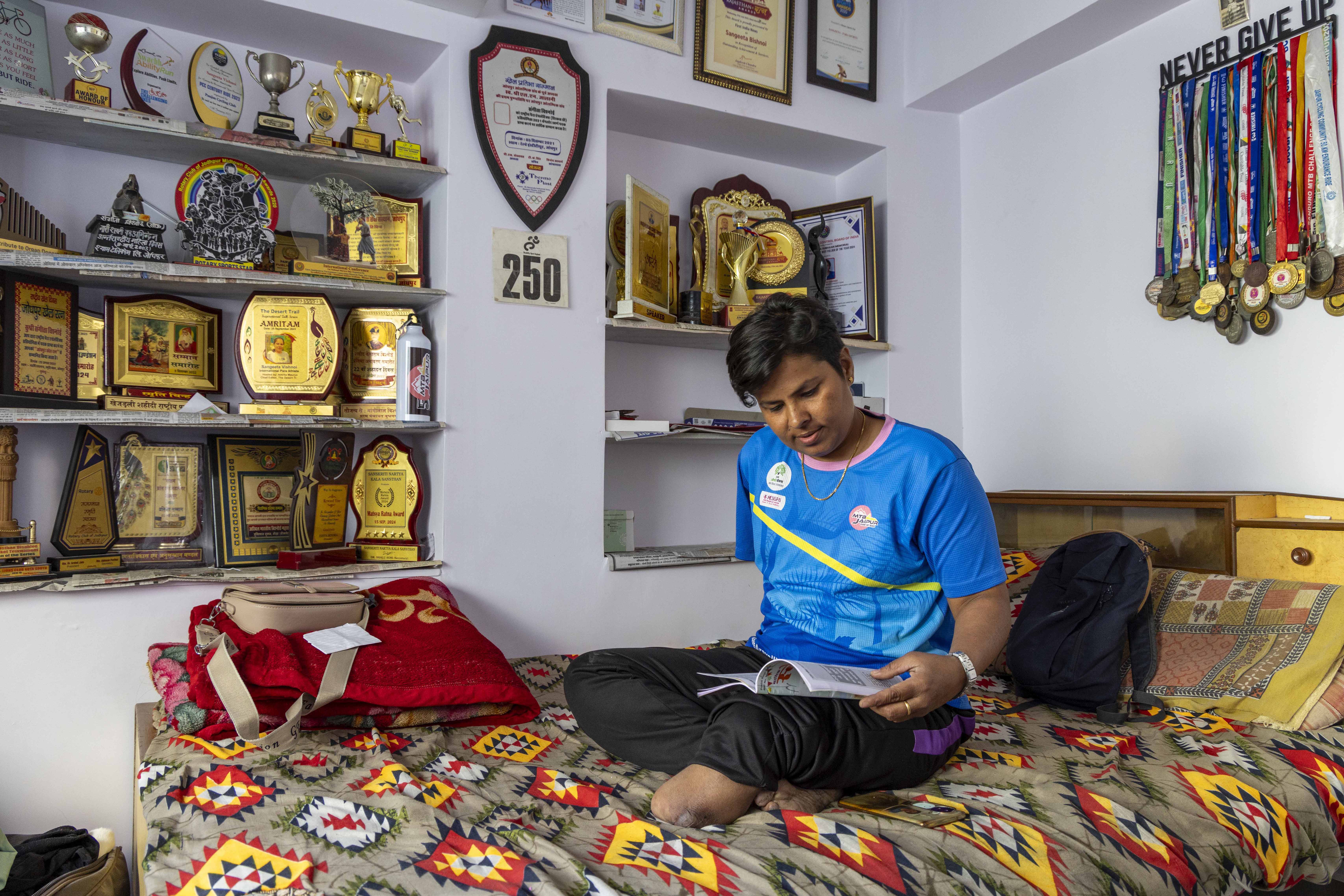 Sangeeta Vishnoi (35) sits cross-legged on her bed, reading a slim book she holds open with her left hand. She has no right hand. She has black, straight hair with a side parting, cut short till it just touches her ears. She wears a dark sky blue T-shirt and black track pants. The bed is in the corner of a room with white walls with a pale lilac tinge. The handloom cotton bedsheet has an ikkat pattern of red, yellow and black on a smoky-grey background. The left side wall has two built-in, three-tiered shelves crammed with plaques, gleaming golden and showing hints of rosewood-coloured wood. There are also several silver and golden cups and trophies. A bunch of silver, golden and bronze medals hang on multi-coloured silken ribbons from a narrow, dark chocolate-coloured, wooden pelmet nailed to the right side wall. Just above the horizontal pelmet are these words carved out in capitals in chocolate-coloured wood: NEVER GIVE UP.