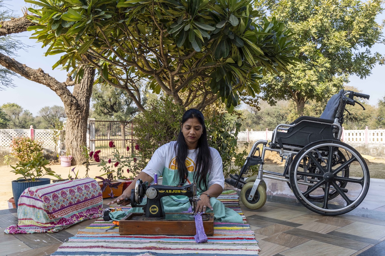 Seema Kanwar (37) sits cross-legged behind a black hand-sewing machine on a cotton rug spread out on a low platform. The platform, which has marble tiles in pale shades of grey and pink, is an extension of the front of the house and is open to the skies. The white rug has thin stripes of pink, blue, maroon, yellow, purple and grey. Seema has long straight black hair that is backcombed, with tresses falling down each shoulder. She wears a white T-shirt that turns into light sky blue from the midriff down, and a matching light sky-blue skirt. Just behind her there is a plumeria tree with green, racquet-shaped leaves with its branches forming an umbrella over her. There is a green shrub next to it and potted plants around the perimeter of the platform. On the platform to the right of her there is a slate-grey motorised wheelchair Seema holds the wheel of the sewing machine with her right hand while her left hand holds a band of grey fabric in place beneath the needle.