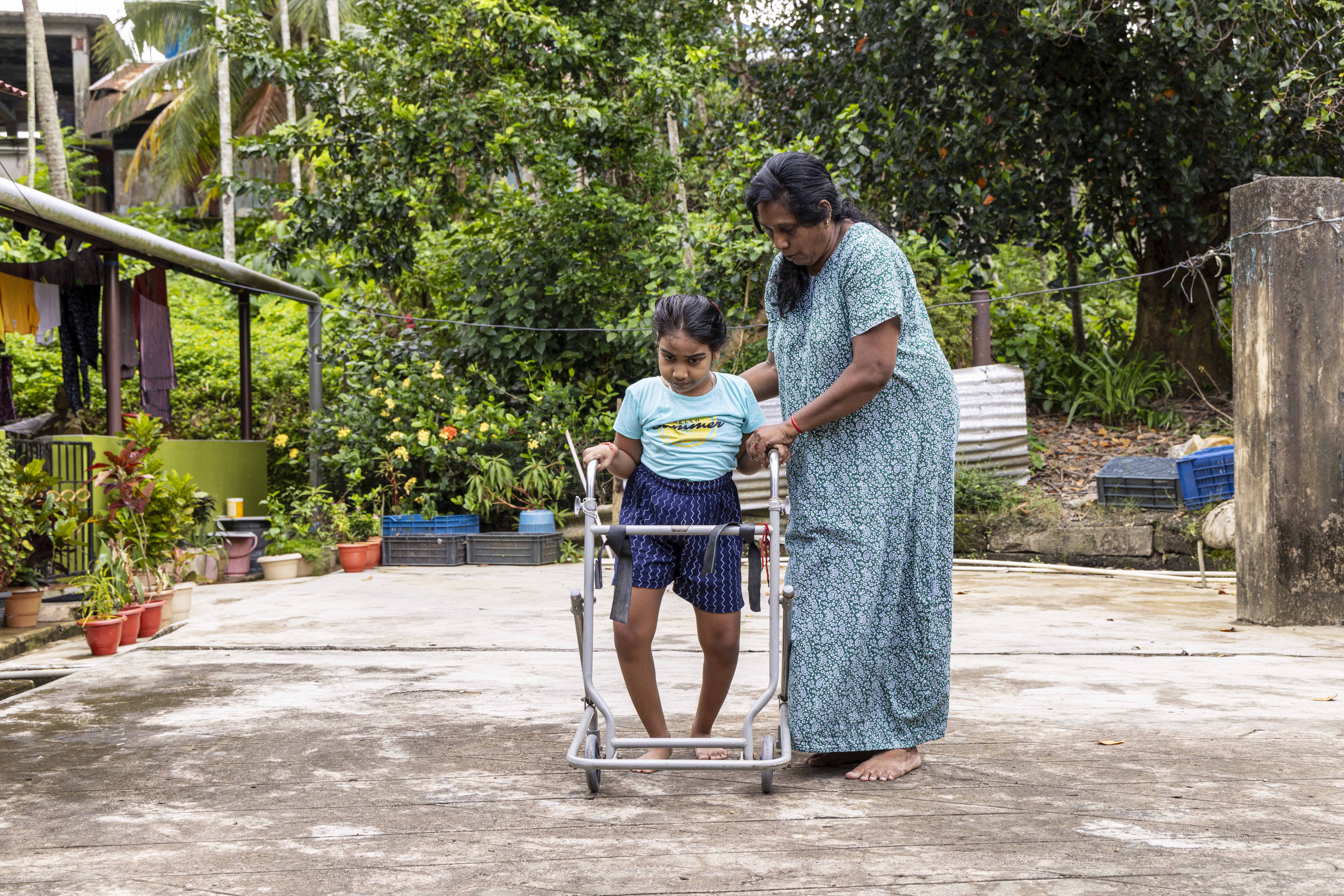 Shaziya Hameed (7) is outdoors, approaching the camera with her aunt Jaygun Bibi (55) who is assisting her walk with the CP walker meant for children with cerebral palsy (CP). Both are barefoot. The whitish ground is paved with cement and the far end of the compound is edged with trees and flowering plants. Shaziya’s hair is tied back in a centre parting. She wears a sky blue T-shirt and navy blue shorts printed with thin, wavy, vertical, parallel lines. The aluminium walker, which rolls on castors, has a handrail resembling a semi-rectangular handlebar. Shaziya stands, placing one hand on either side of the handrail. Jaygun, wearing a half-sleeved leaf-green nightie covered with tiny white motifs, stands to the right. She is supporting Shaziya’s back with her right hand while gripping Shaziya’s left hand with her left.