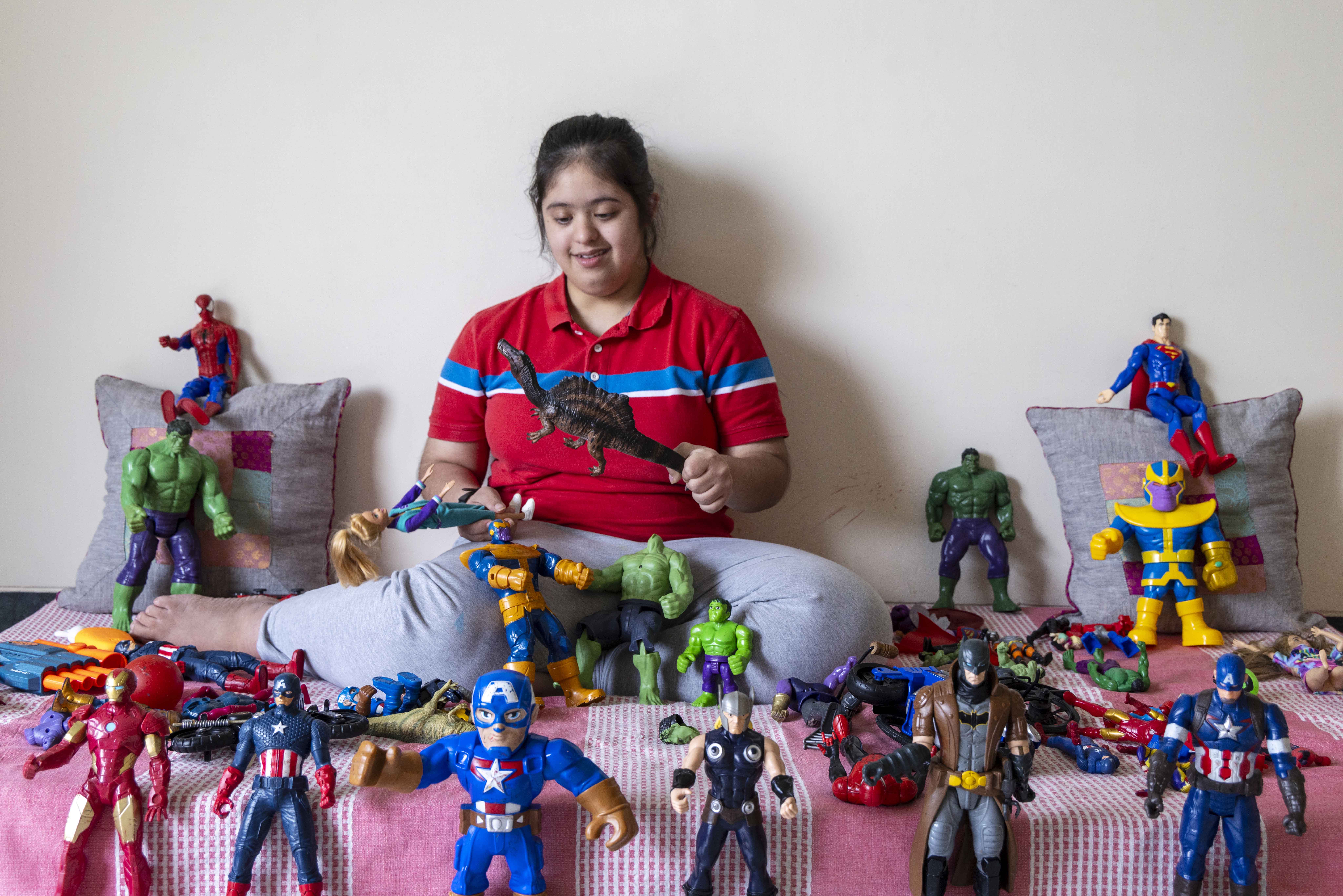 Shreya Dhillon (15) sits on a bed with legs folded to one side. Her black hair is combed back and tied. She wears pale blue jeans and a red, half-sleeved, knitted shirt with a single, horizontal, broad, dark sky-blue stripe edged in white running across and above her chest. She is surrounded by dozens of action figure toys, of Superman, Spiderman, Batman, Captain America, Hulk and other heroes, in every shade of the colour spectrum. Some stand upright in a row in front of her and others are clustered around her or on her lap. Action figures are also atop two upright, square, light grey cushions on either side of her. She holds a chocolate brown toy dinosaur in her left hand and a Barbie doll in a blue outfit in her right. The bedsheet has broad panels of light pink and white.