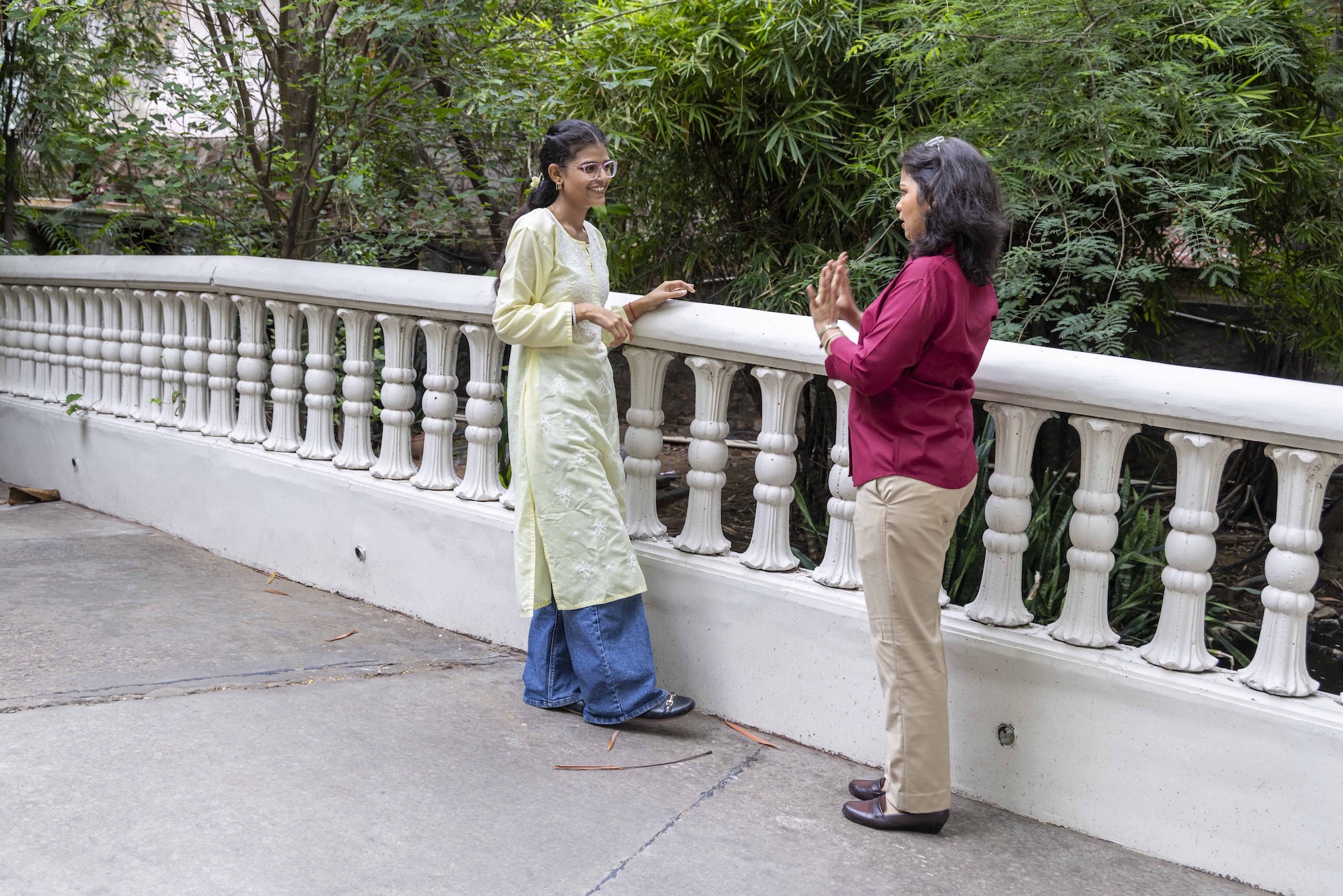Simran Kumari (22) stands outdoors facing another young woman next to a decorative concrete railing painted white. The railing has a cylindrical moulded handrail and stout, ornamental balusters. Simran wears blue jeans that peep out from beneath a calf-level, pale yellow, full-sleeved, round-necked kurta with white chikankari embroidery on the front. She wears glasses with a silvery plastic rectangular frame and her hair is tied back with a centre parting. The woman standing opposite her wears a blood-red full-sleeved shirt over beige trousers. She shows her raised palms to Simran who is smiling at her.