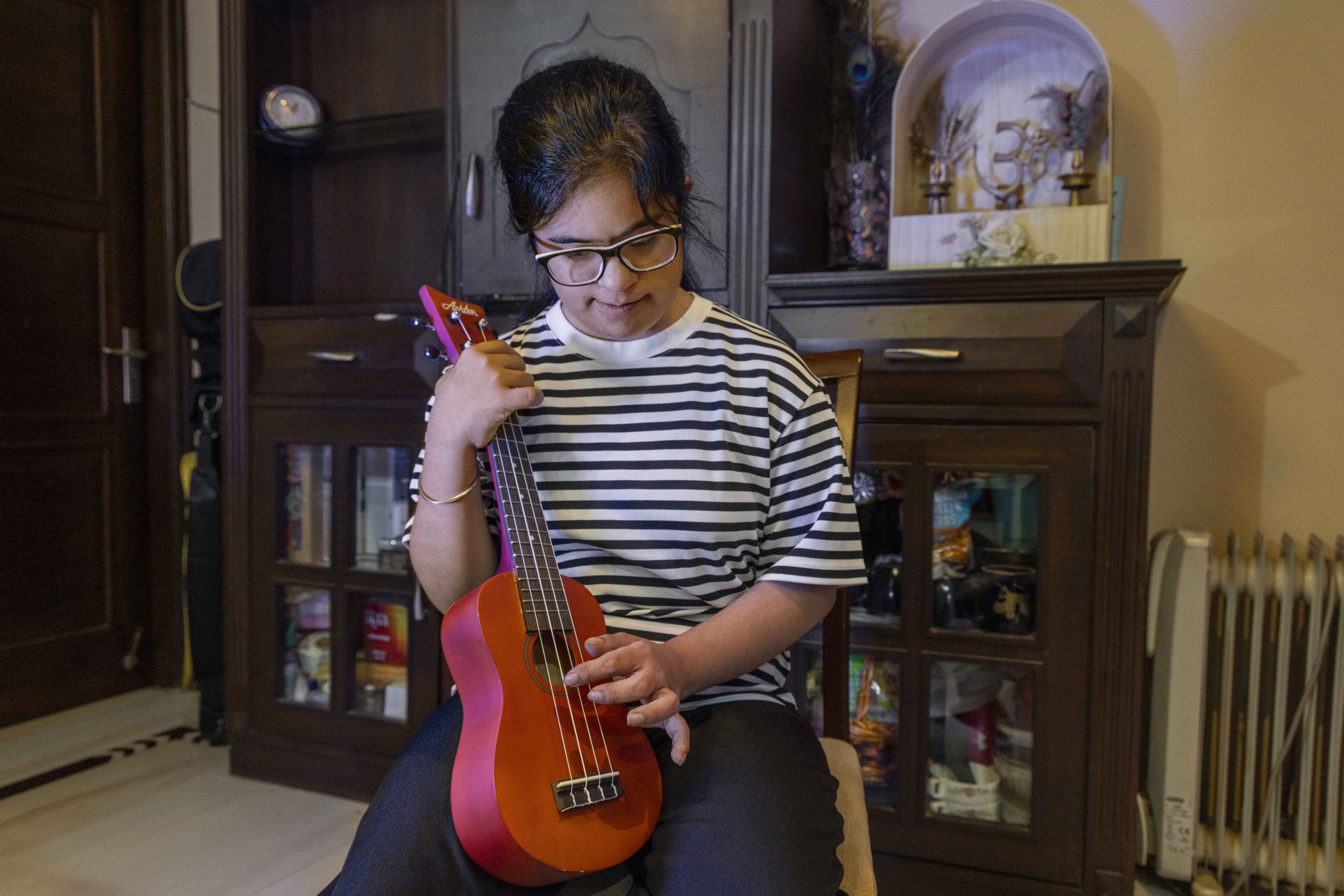 Simran Wadhwa (21) sits on a wooden chair, holding a toy guitar. She wears specs with black frames and her straight hair is combed back and tied. She is dressed in black pants and a T-shirt with horizontal black and white stripes. The dark pink guitar has four strings. Simran has wrapped her right hand around the head of the guitar and she is plucking a string on the body of the guitar with her left hand. Behind her there is a chocolate brown wooden showcase with framed glass panes and two top drawers.
