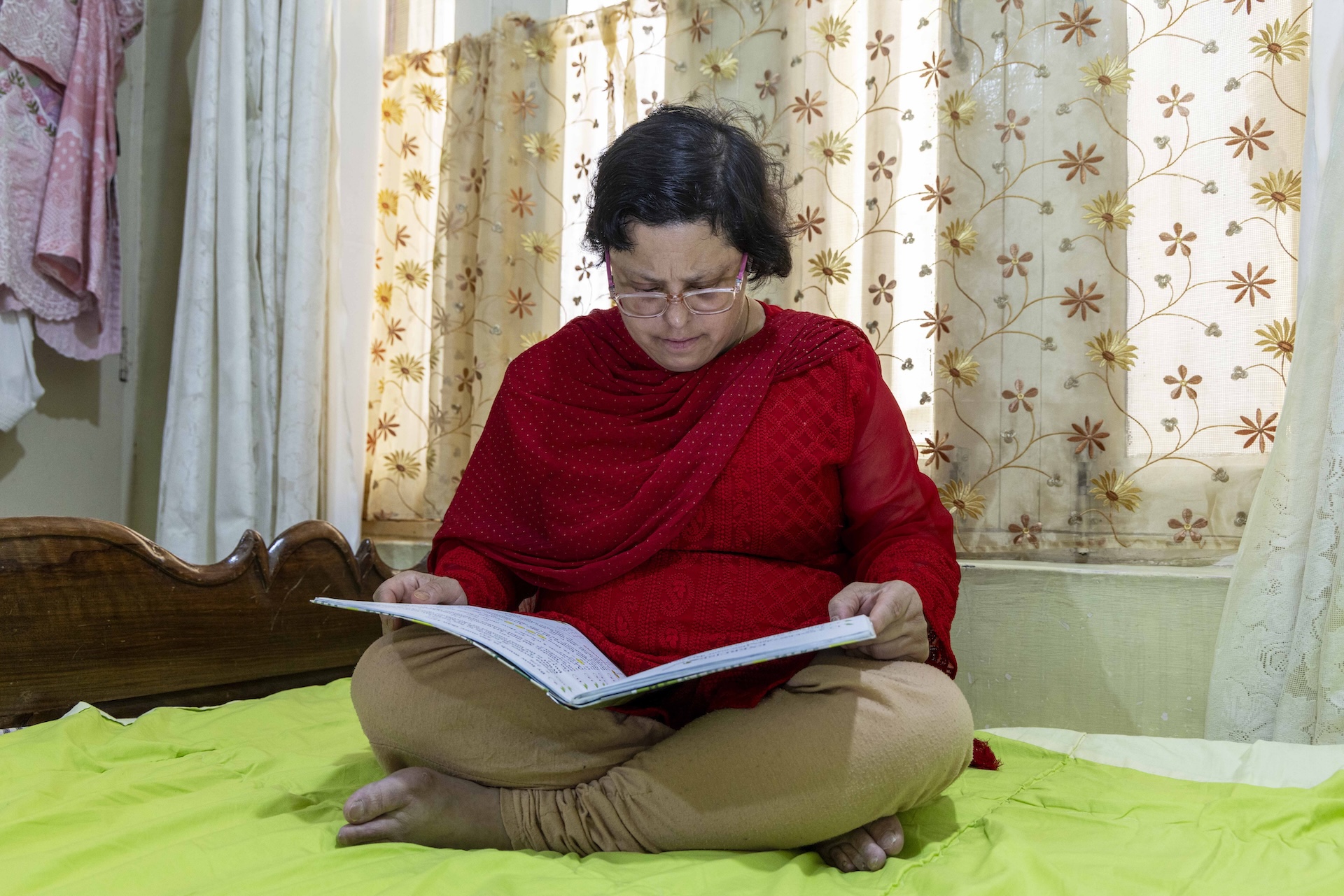 Sofia Qadir Tak (38) sits cross-legged on a wooden bed. She wears specs with a pinkish plastic rectangular frame. Her bobbed hair has a side parting. She is dressed in a blood-red full-sleeved kameez, a matching dupatta over her shoulders, and a beige churidar. She sits on a light green sheet covering the mattress. A large thin book is open on her lap and she is looking down at it. The drawn window curtain behind her is cream with brown floral motifs.