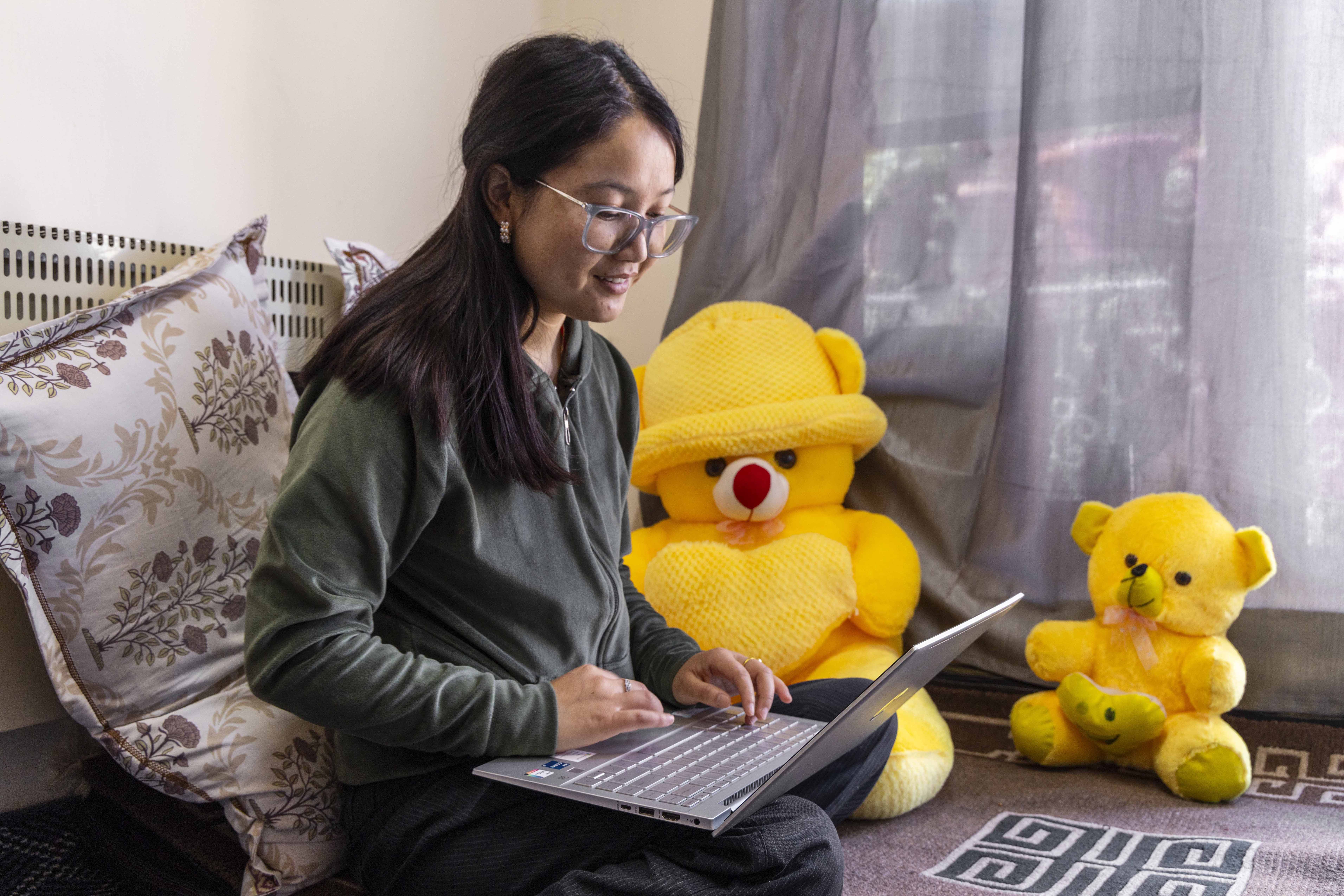 Sonam Yangchan (26) sits on a pale chocolate-coloured carpet with legs folded at the knees and tucked beneath her. She has straight hair with a centre parting, which falls to just below her shoulders. She wears glasses with a light-coloured rectangular frame. She is dressed in ash grey corduroy trousers and olive-green sweatshirt zipped up at the front. A silvery grey open laptop rests on her lap and she is looking at the screen with fingers on the keyboard. On the carpet next to her are two sunny yellow teddy bears, one small and one large. She leans against a pillow encased in a pale mocha cover printed with a brown floral pattern. Next to the pale mocha wall behind her there is a smoky grey door curtain.