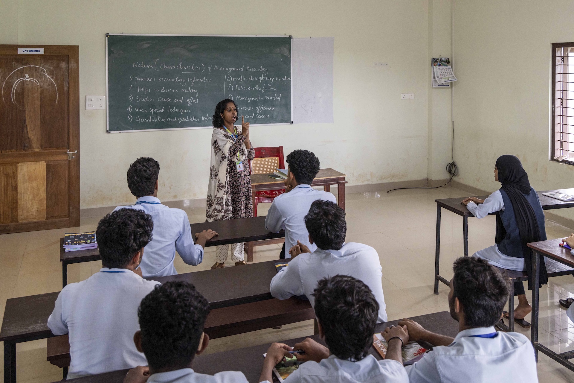 Subitha MC (34) stands facing the camera in a classroom with cream walls and beige floor. In front of her there are rows of chocolate brown wooden desks and benches occupied by teenaged college students with their backs to the camera. Subitha has curly black hair that touches the base of her neck. She wears a cream salwar with small chocolate-brown polka dots and a full-sleeved kameez in a busy floral design of chocolate brown and red. A cream dupatta is draped over her back and chest. Her mouth is partly open and both arms are raised to shoulder level with hands clasped. Her right forefinger points upwards and the other fingers are closed. On the blackboard behind her there are words written in white chalk in point form numbered 1 to 5 to the left and 6 to 10 to the right. The points are the title “Nature (Characteristics) of Management Accounting”. The college uniform is light sky blue and black: the boys wear blue shirt and black trousers and the girls wears blue kameez with a black vest, and black salwar.